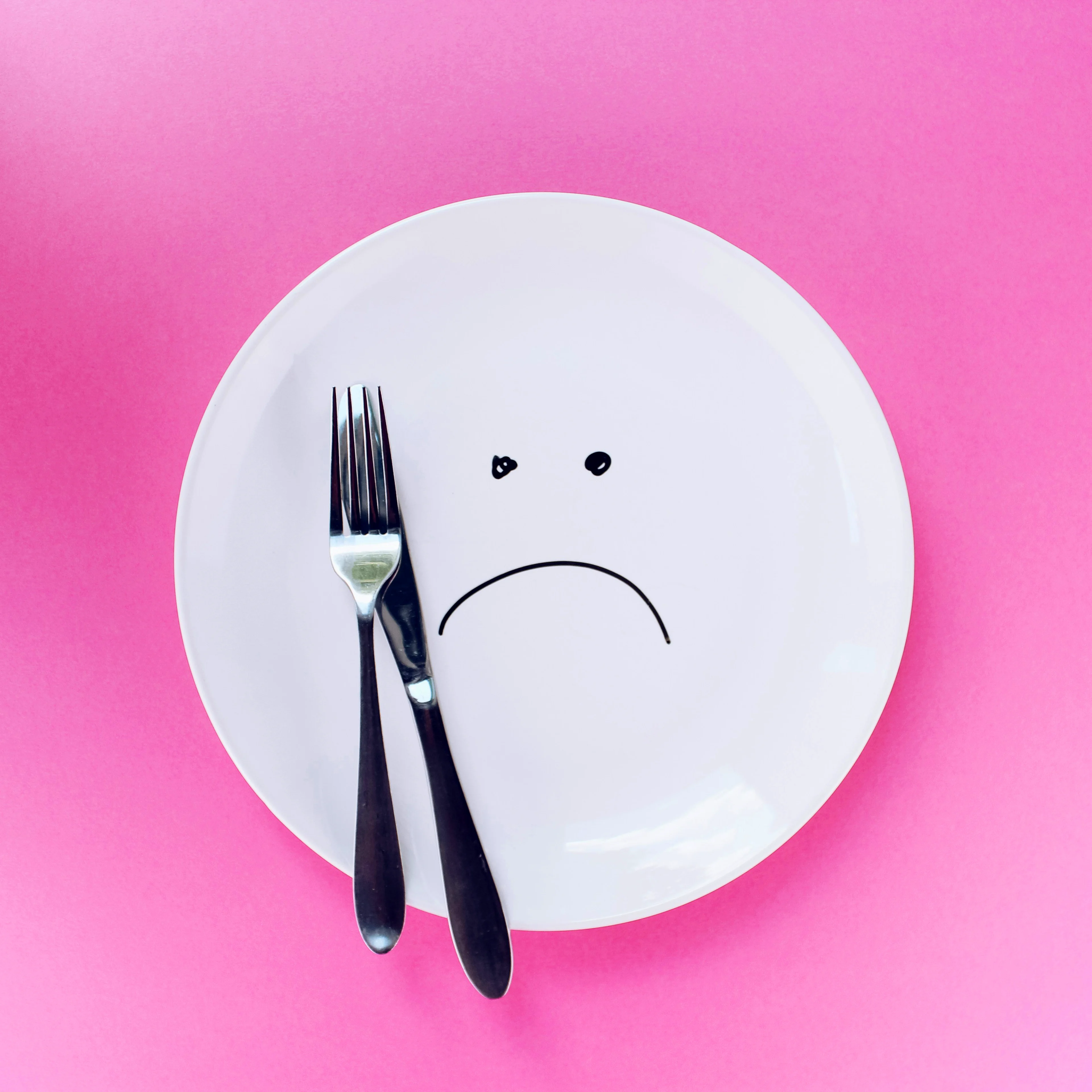 A white plate with a drawn sad face sits on a pink background, with a silver fork and knife resting on it.