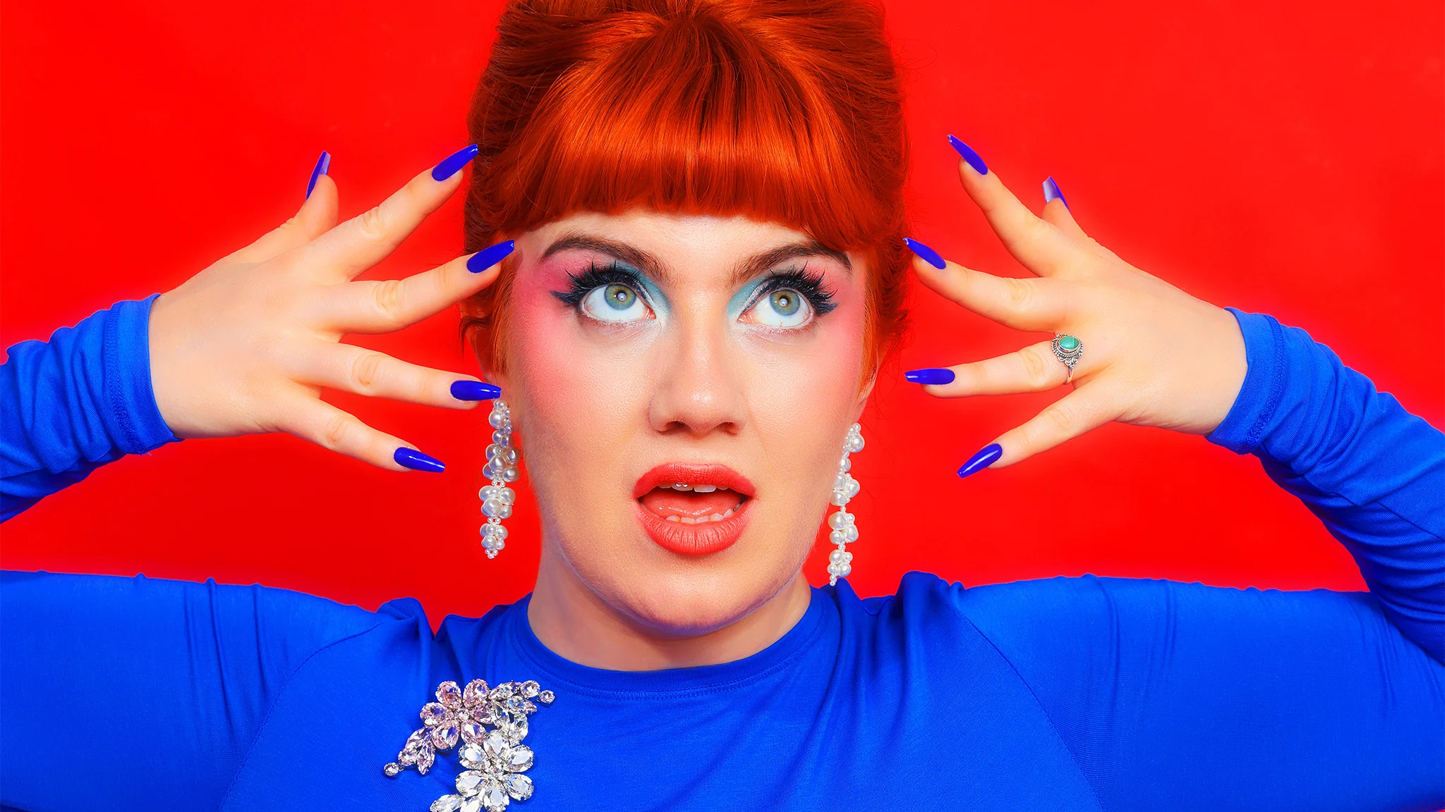 A performer with red hair, bold makeup, blue nails, and a blue outfit poses dramatically against a red background.