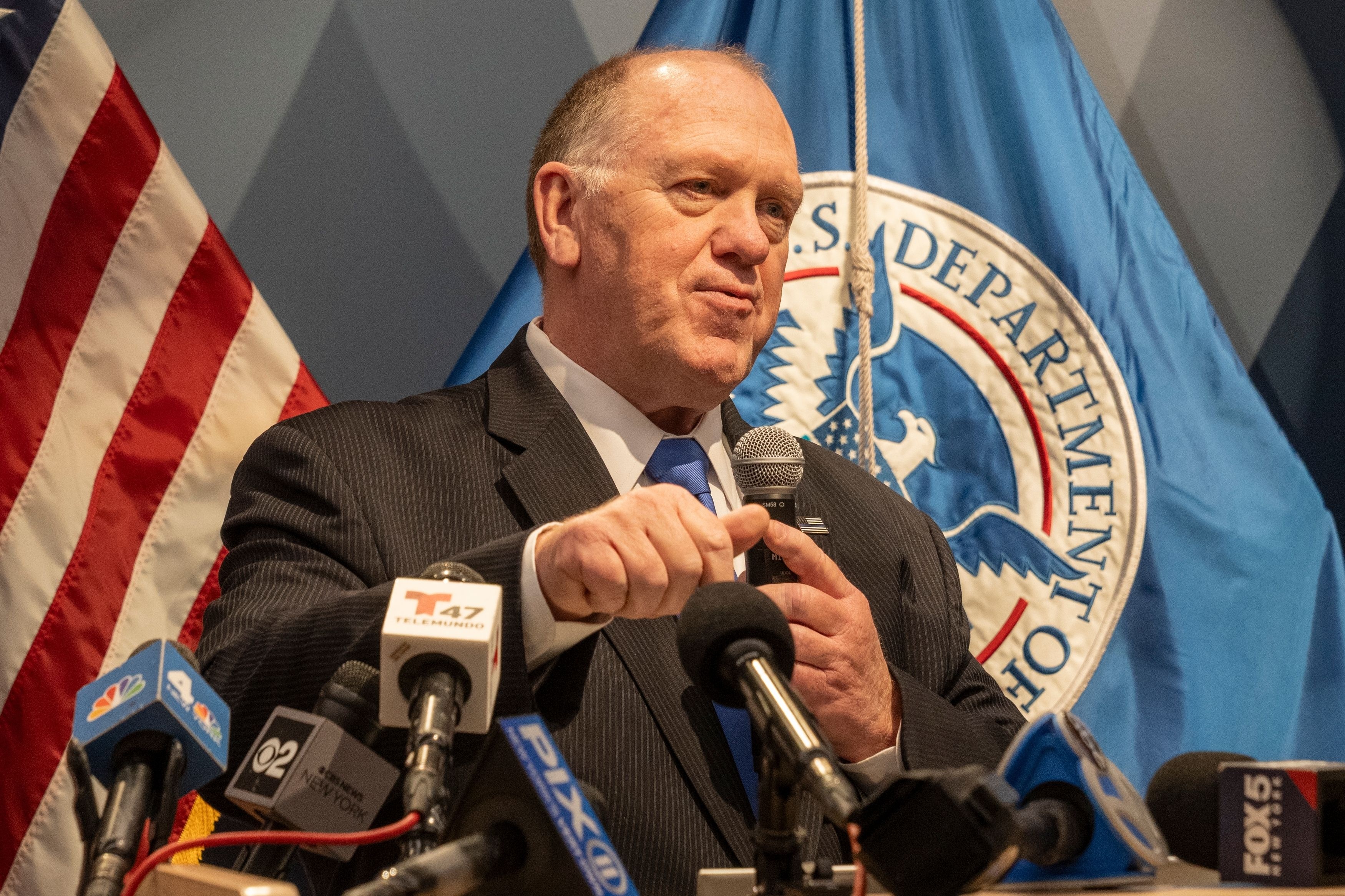 A man in a dark suit speaks at a podium filled with microphones.