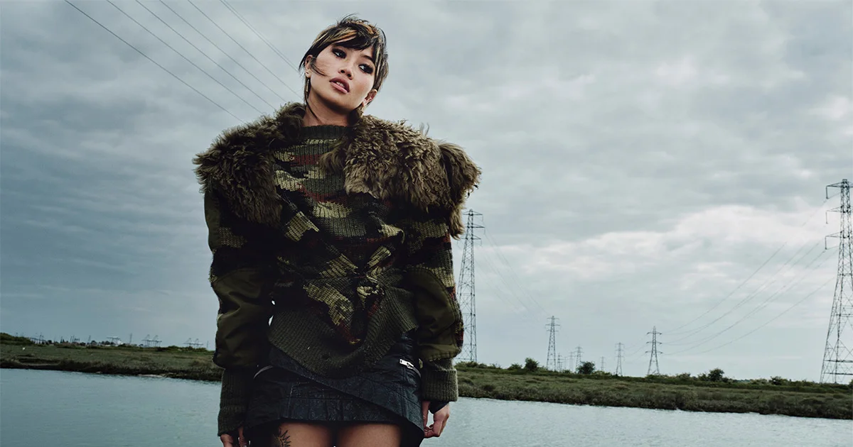 Woman in camo knit sweater with fur trim and mini skirt stands near a river with power lines under a cloudy sky.