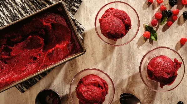 Homemade berry sorbet in glass bowls with fresh berries scattered nearby and a pan of sorbet on a textured cloth.
