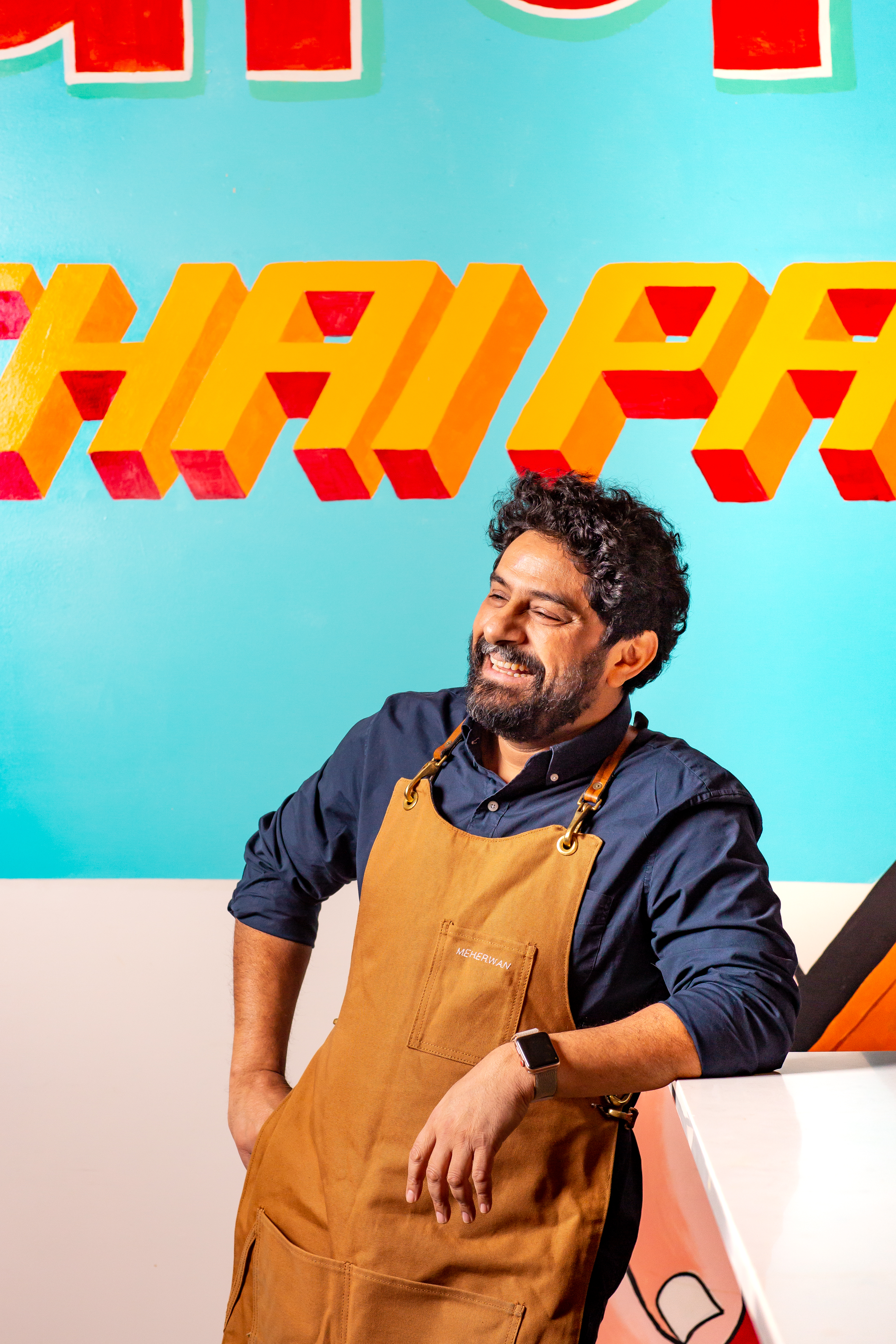 A smiling bearded man named Meherwan wears a tan apron over a navy shirt, posed against a colorful teal mural.