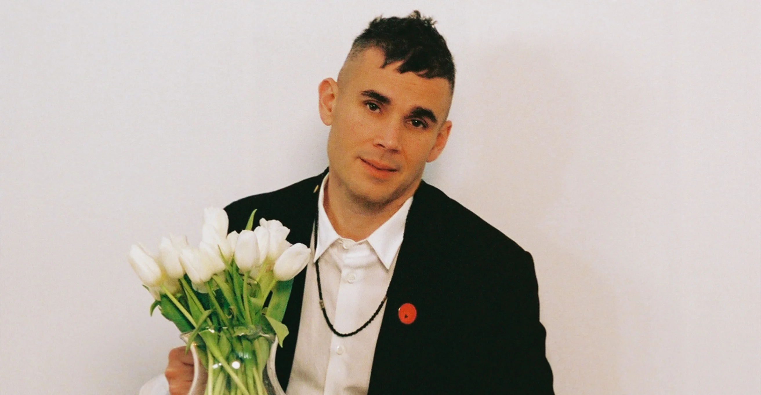 Rostam in a black blazer and white shirt holds a vase of white tulips, wearing a red pin and bead necklace.