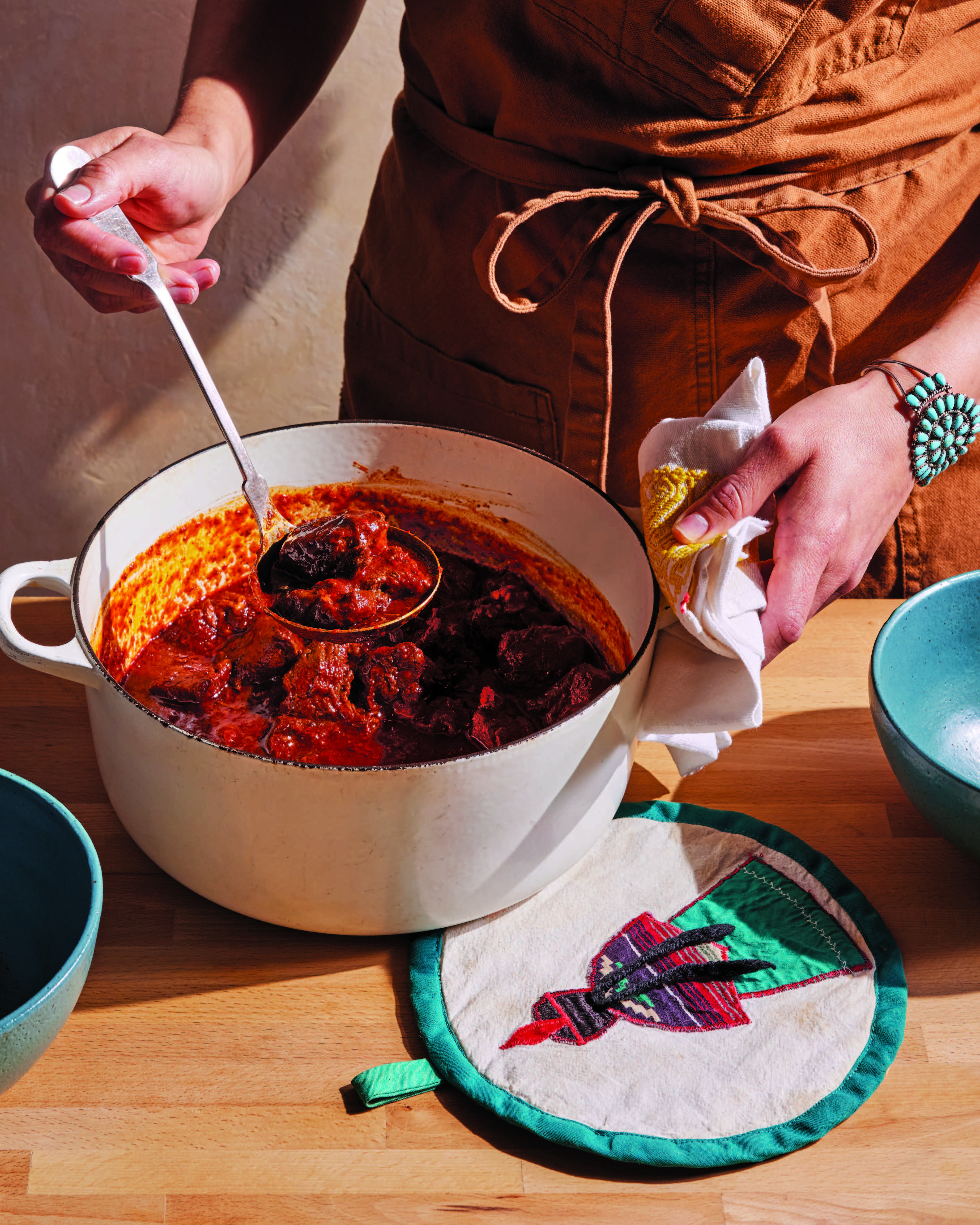 Person in brown apron stirring red meat stew in white Dutch oven, wearing turquoise bracelet, with decorative potholder nearby.