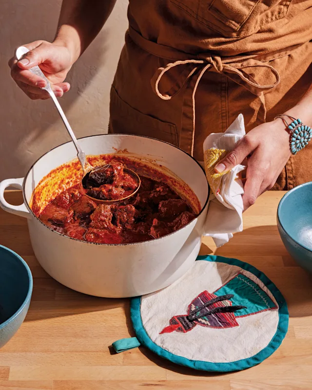 Person in brown apron stirring red meat stew in white Dutch oven, wearing turquoise bracelet, with decorative potholder nearby.