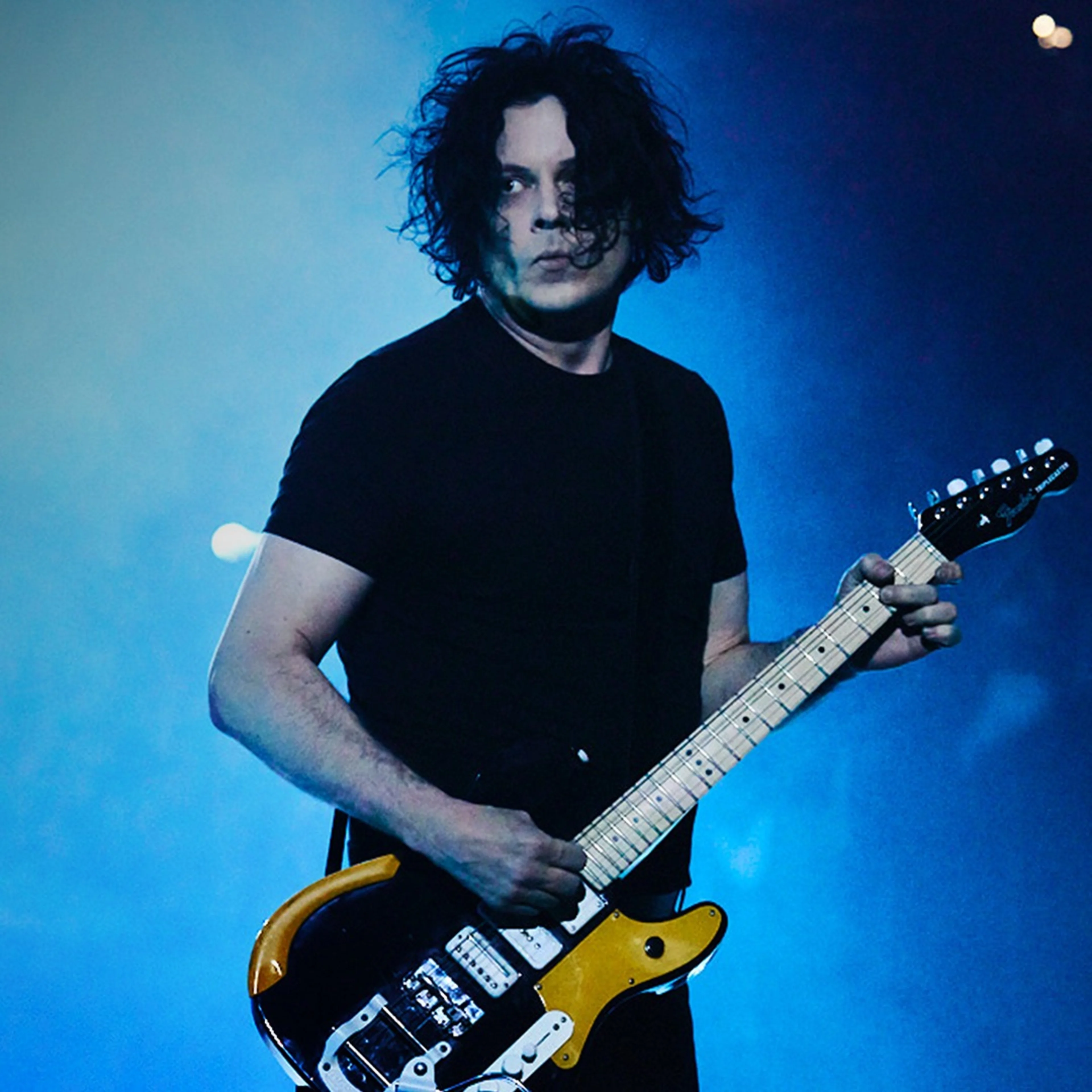 A male guitarist with dark curly hair plays a yellow and black electric guitar on stage, lit by blue lighting.