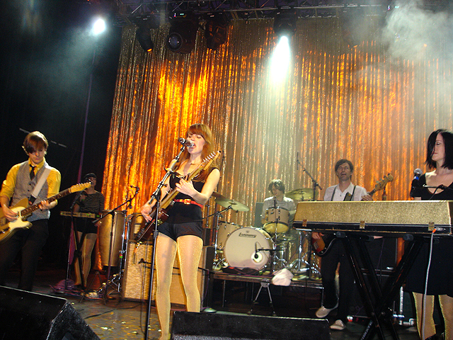 Band performing on stage with gold sequin backdrop, female singer at microphone, musicians playing various instruments.