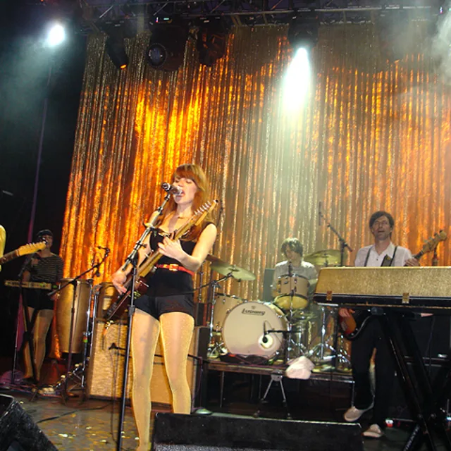 Band performing on stage with gold sequin backdrop, female singer at microphone, musicians playing various instruments.