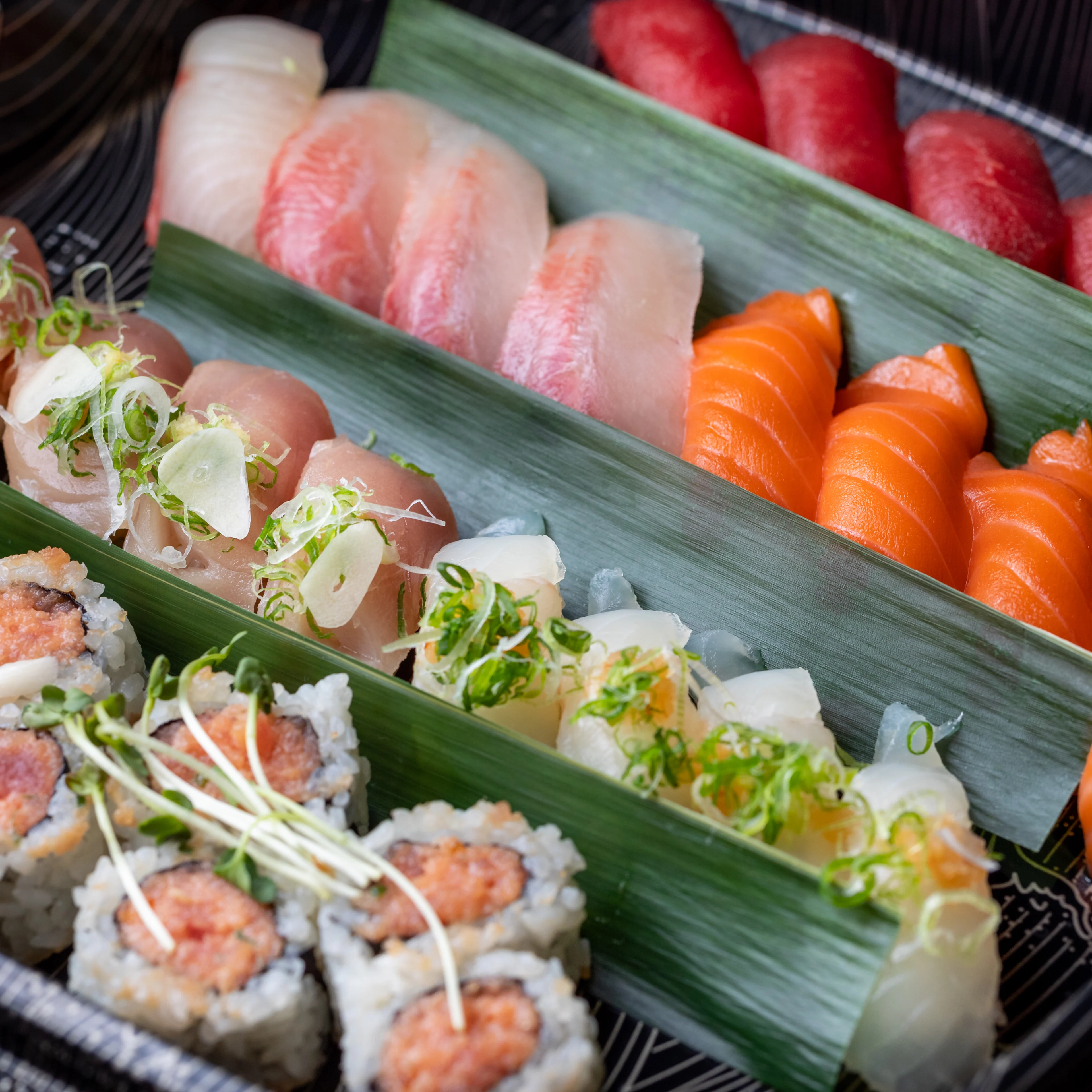 Assorted sushi platter with salmon, tuna and white fish nigiri, maki rolls, and garnishes in a black container.