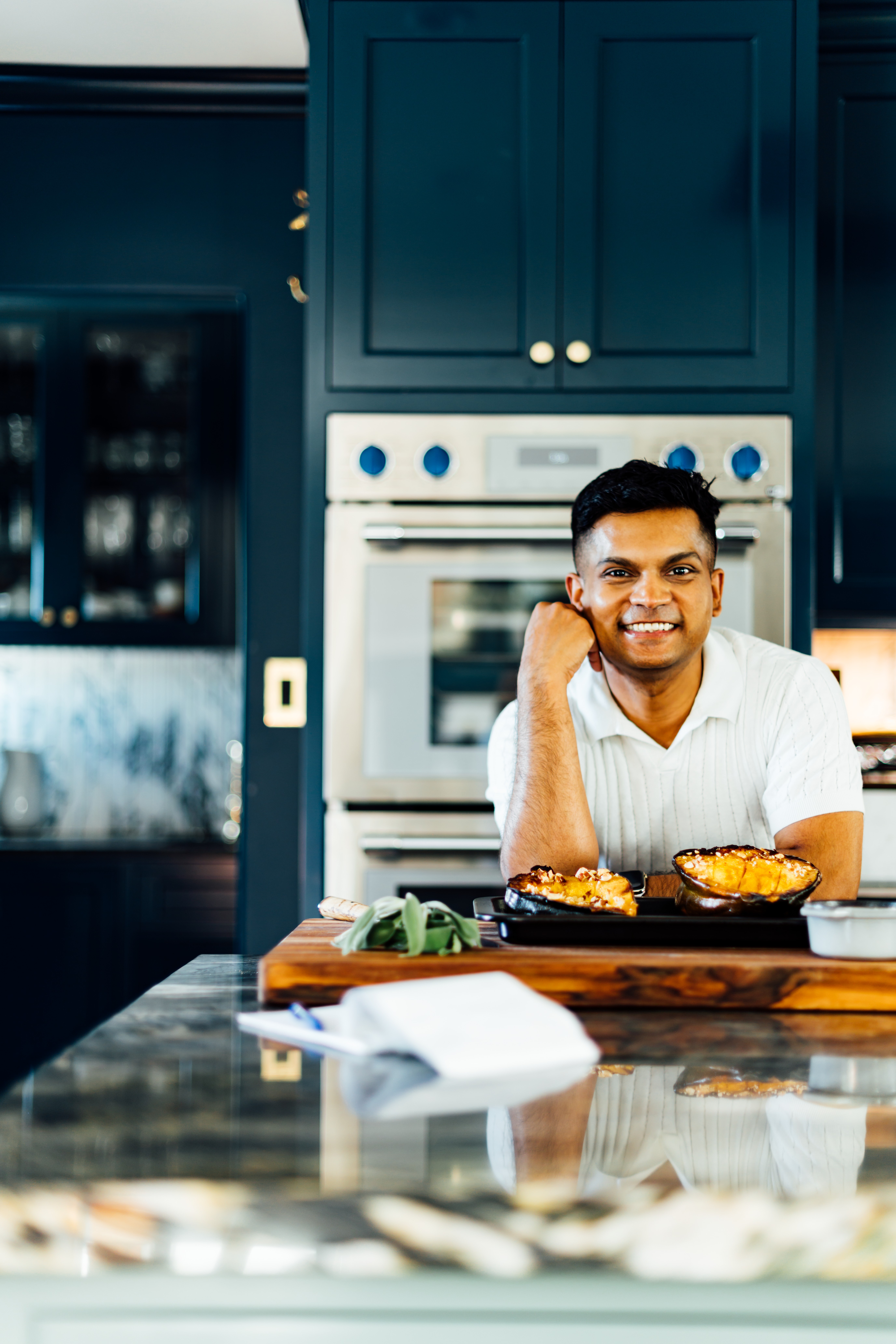 A smiling man in a white shirt leans on a kitchen island with roasted dishes, surrounded by navy blue cabinetry and a double oven.