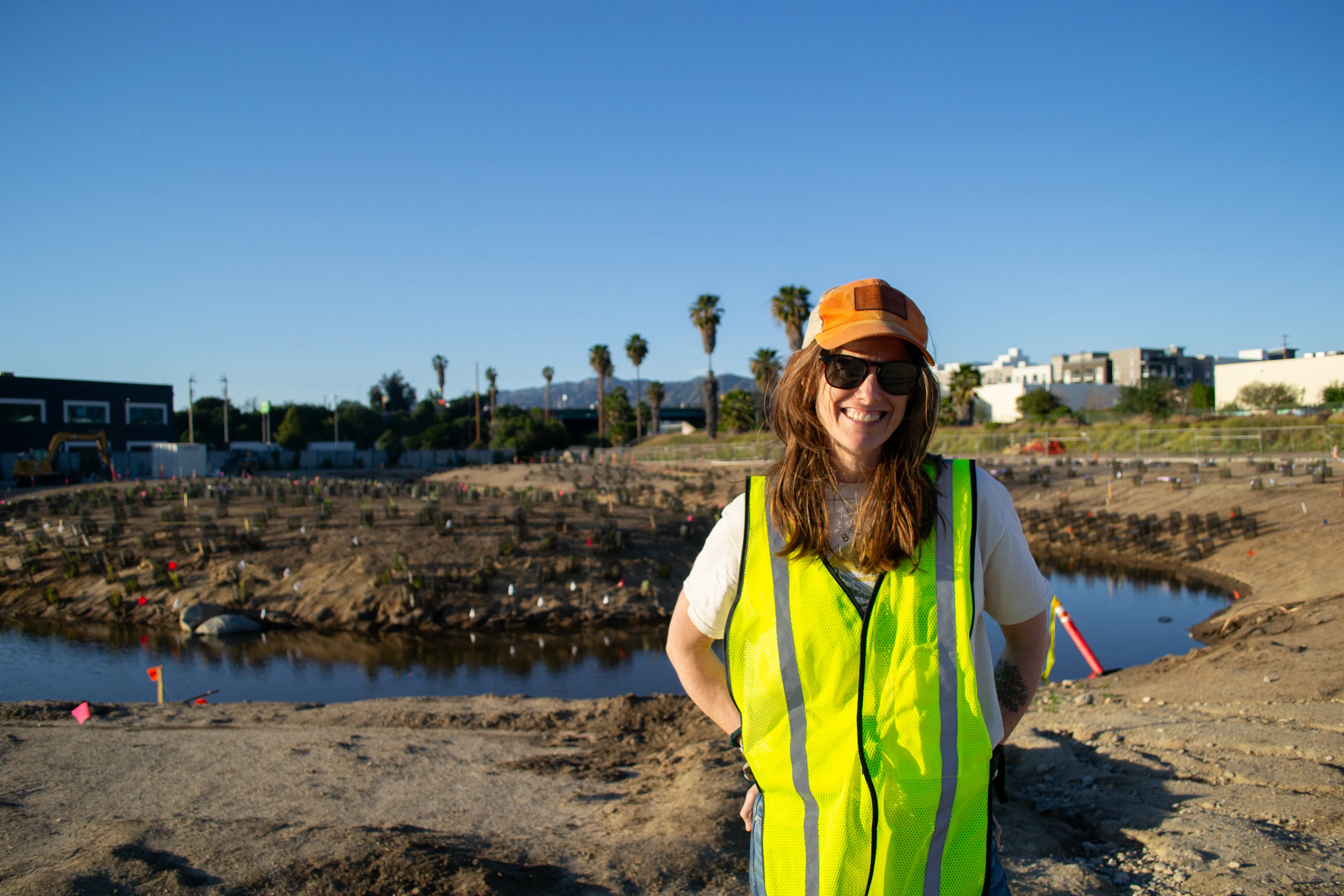 A woman in a neon construction vest smiles in front of the Bowtie Wetland Demonstration site. You can see the winding waterway starting to form behind her.