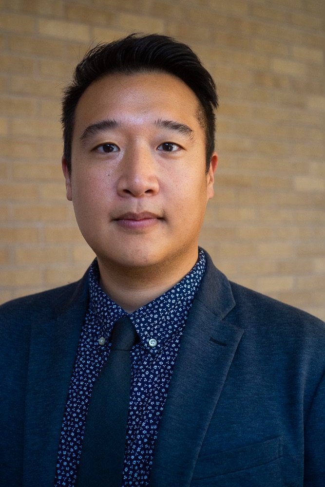 A young man in a gray blazer and navy patterned shirt poses in front of a brick wall.
