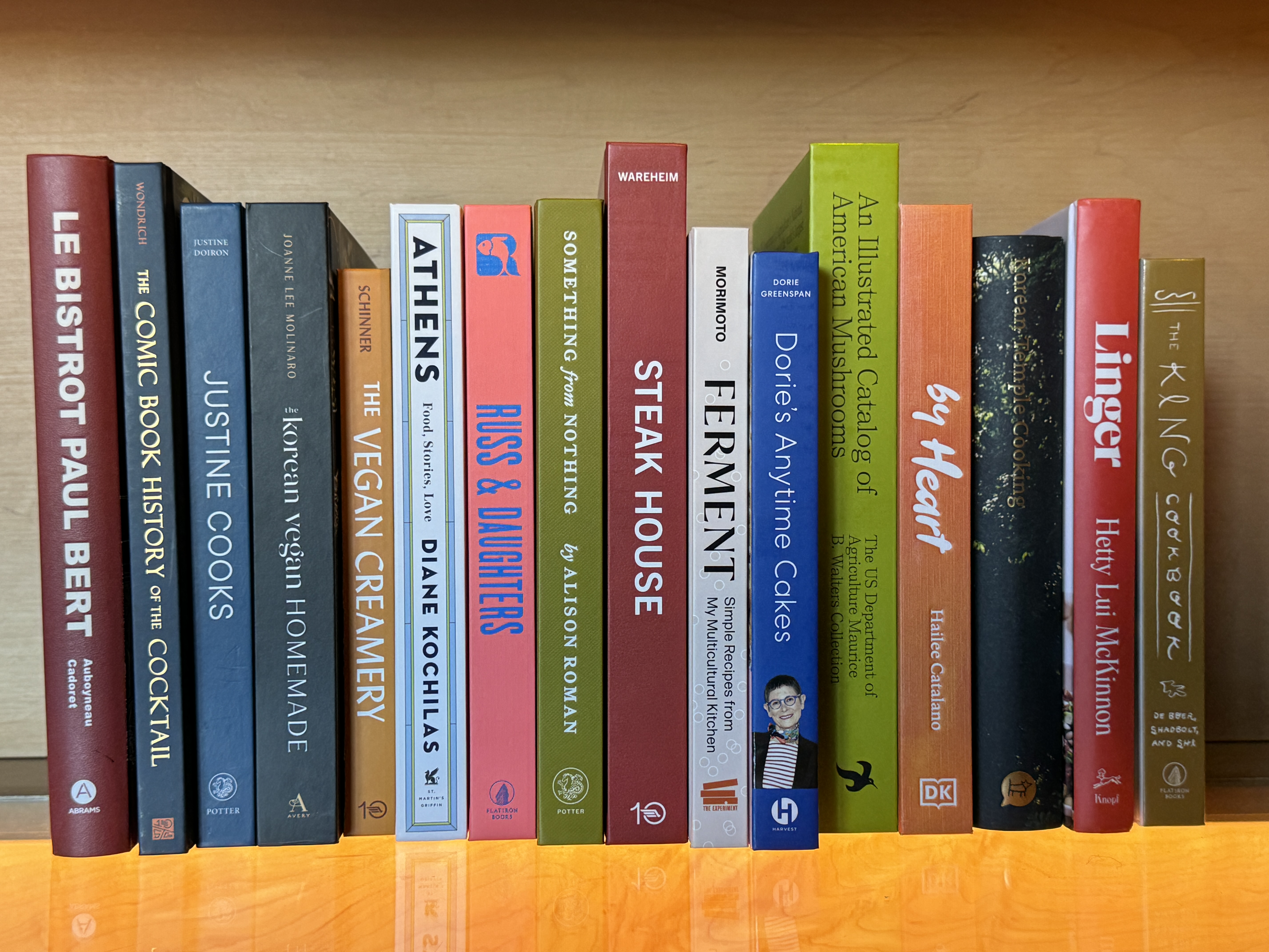 Row of colorful books on a wooden shelf including titles by Diane Kochilas, Alison Roman, and cookbooks on various cuisines.