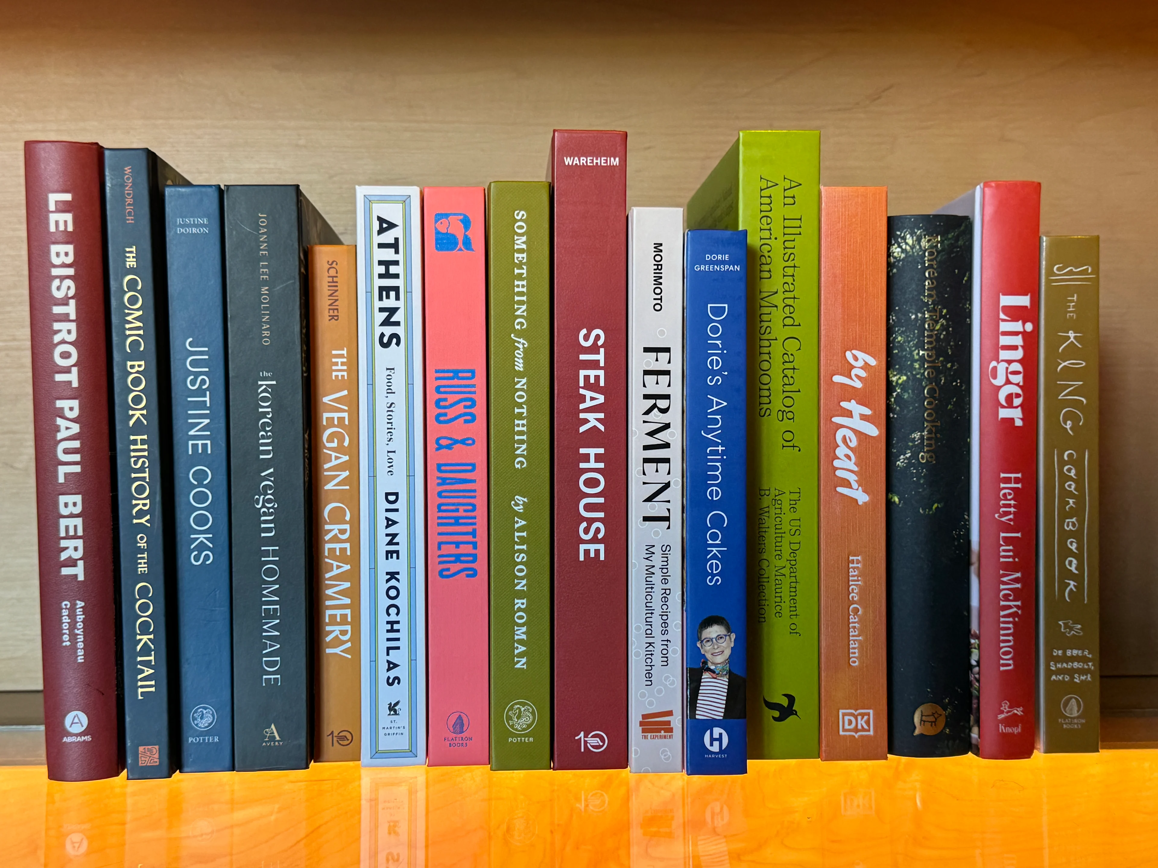 Row of colorful books on a wooden shelf including titles by Diane Kochilas, Alison Roman, and cookbooks on various cuisines.