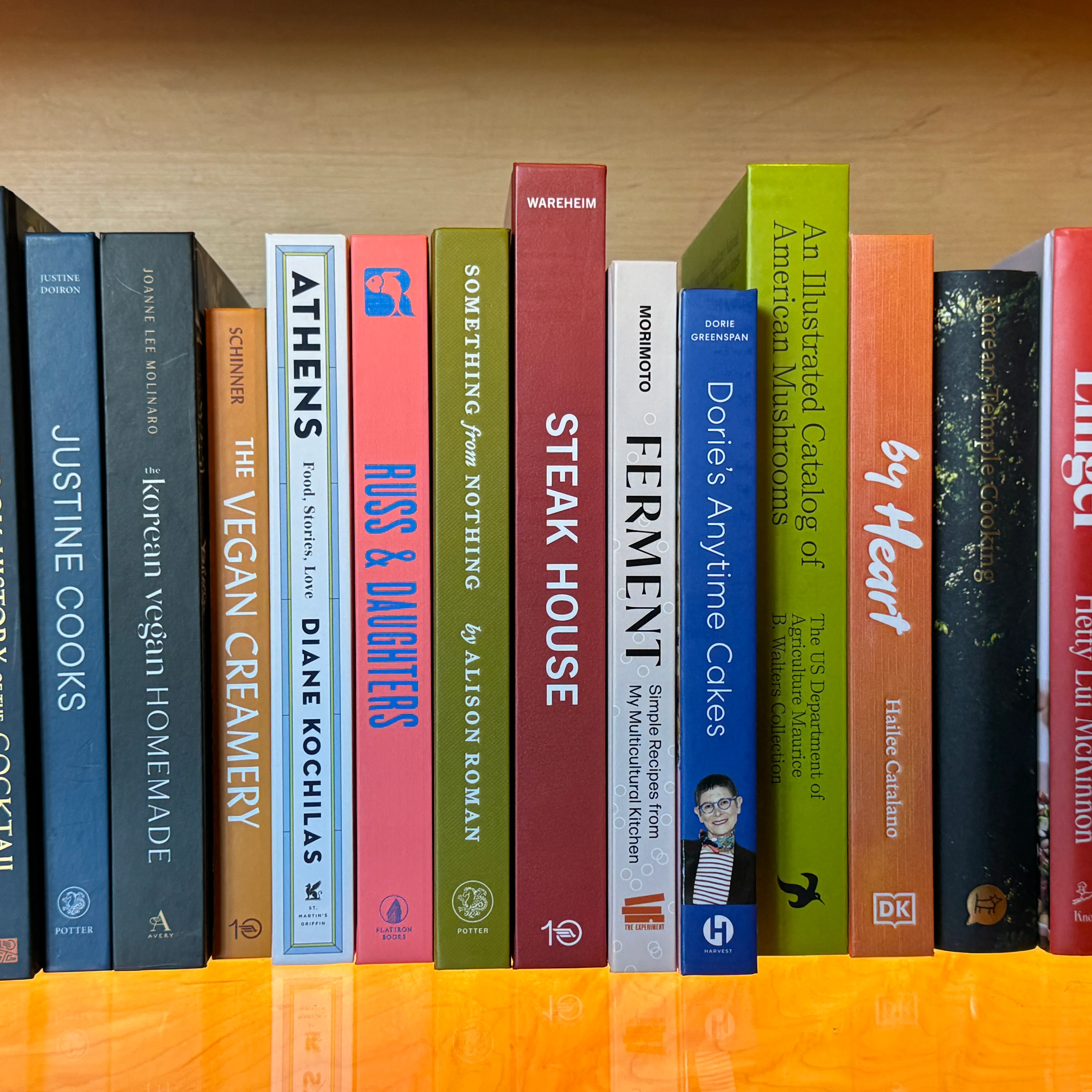 Row of colorful books on a wooden shelf including titles by Diane Kochilas, Alison Roman, and cookbooks on various cuisines.