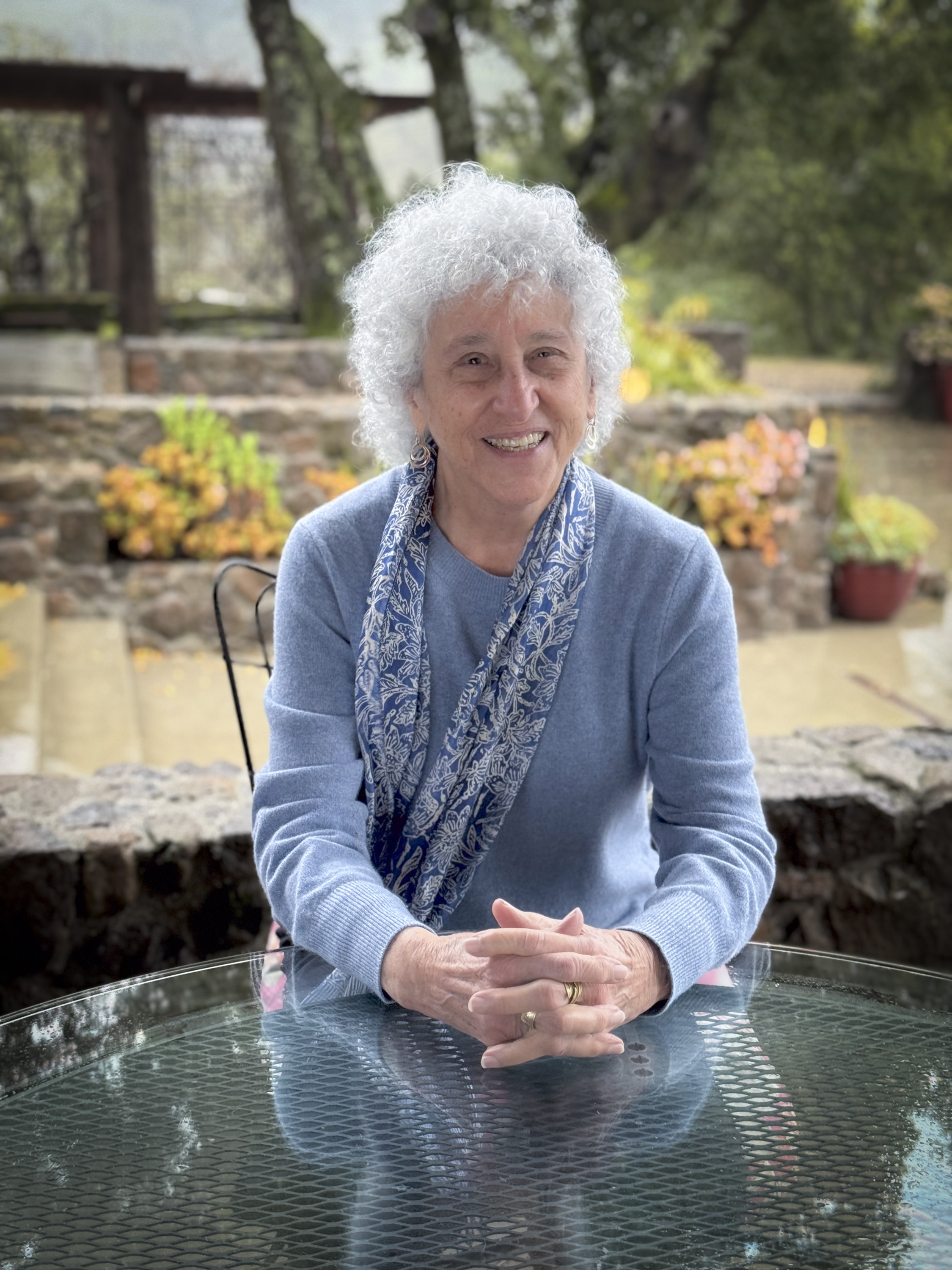 An older woman with white curly hair smiles at a glass patio table, wearing a blue sweater and floral scarf, garden behind her.