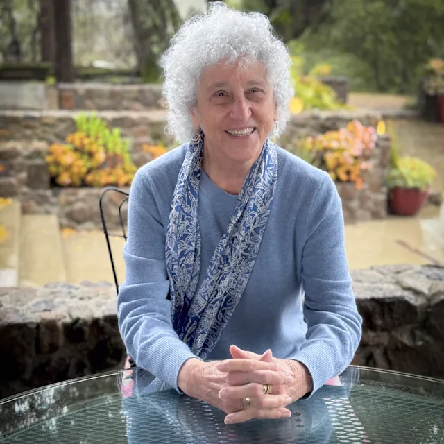 An older woman with white curly hair smiles at a glass patio table, wearing a blue sweater and floral scarf, garden behind her.