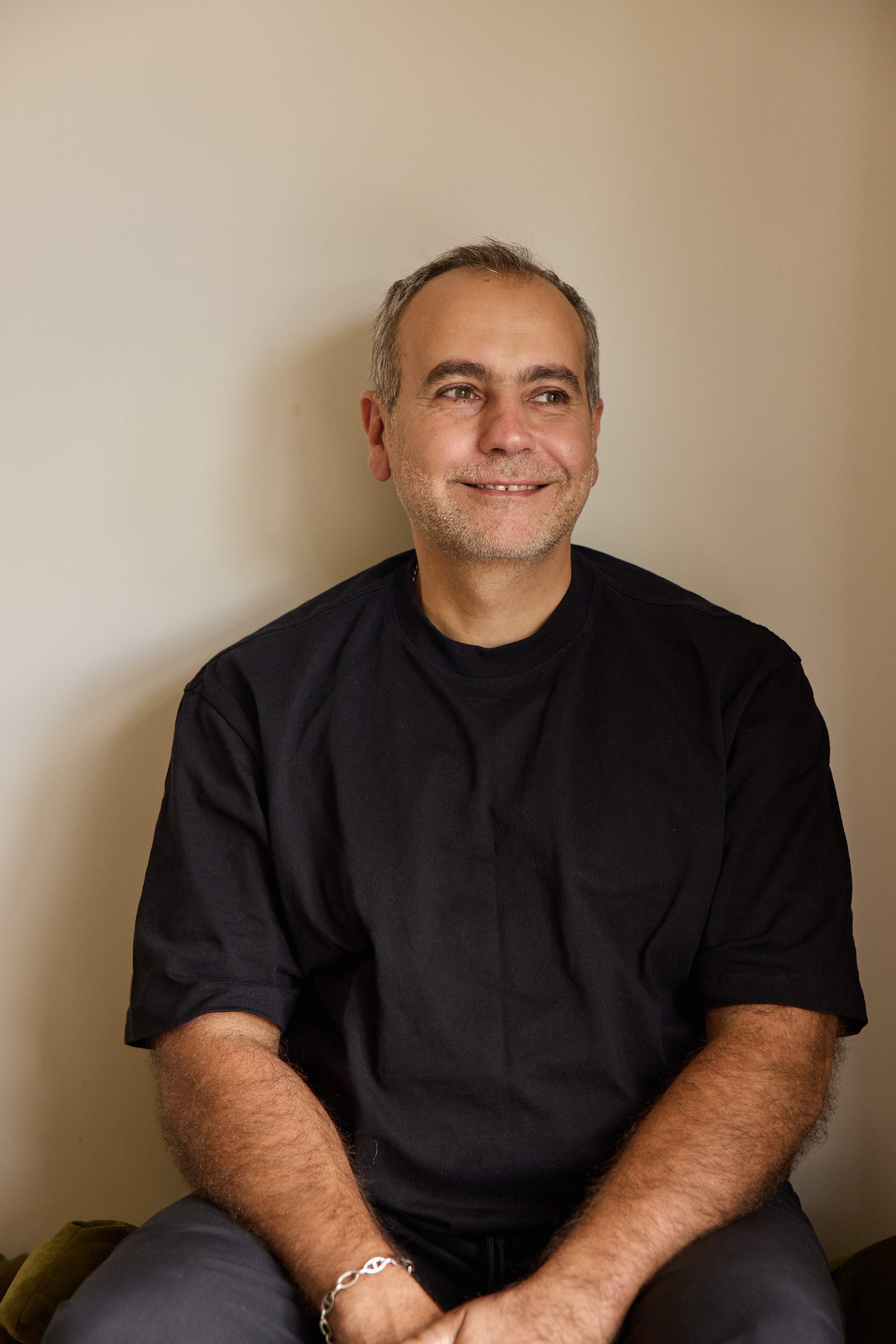 A middle-aged man with gray hair smiles warmly, wearing a black t-shirt, seated against a beige wall.