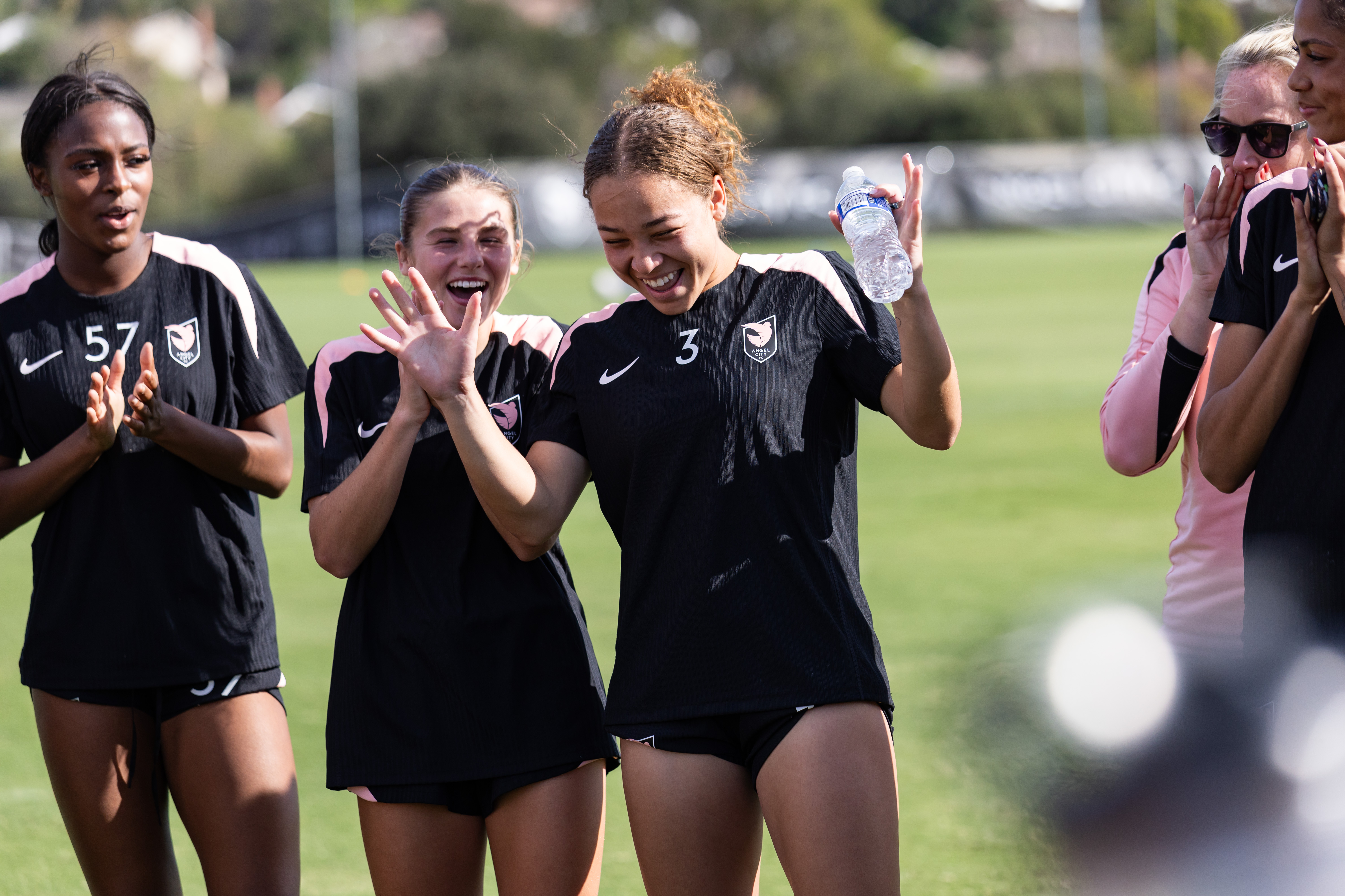 An Angel City FC player in uniform celebrates with her teammates while holding a water bottle after a scrimmage.
