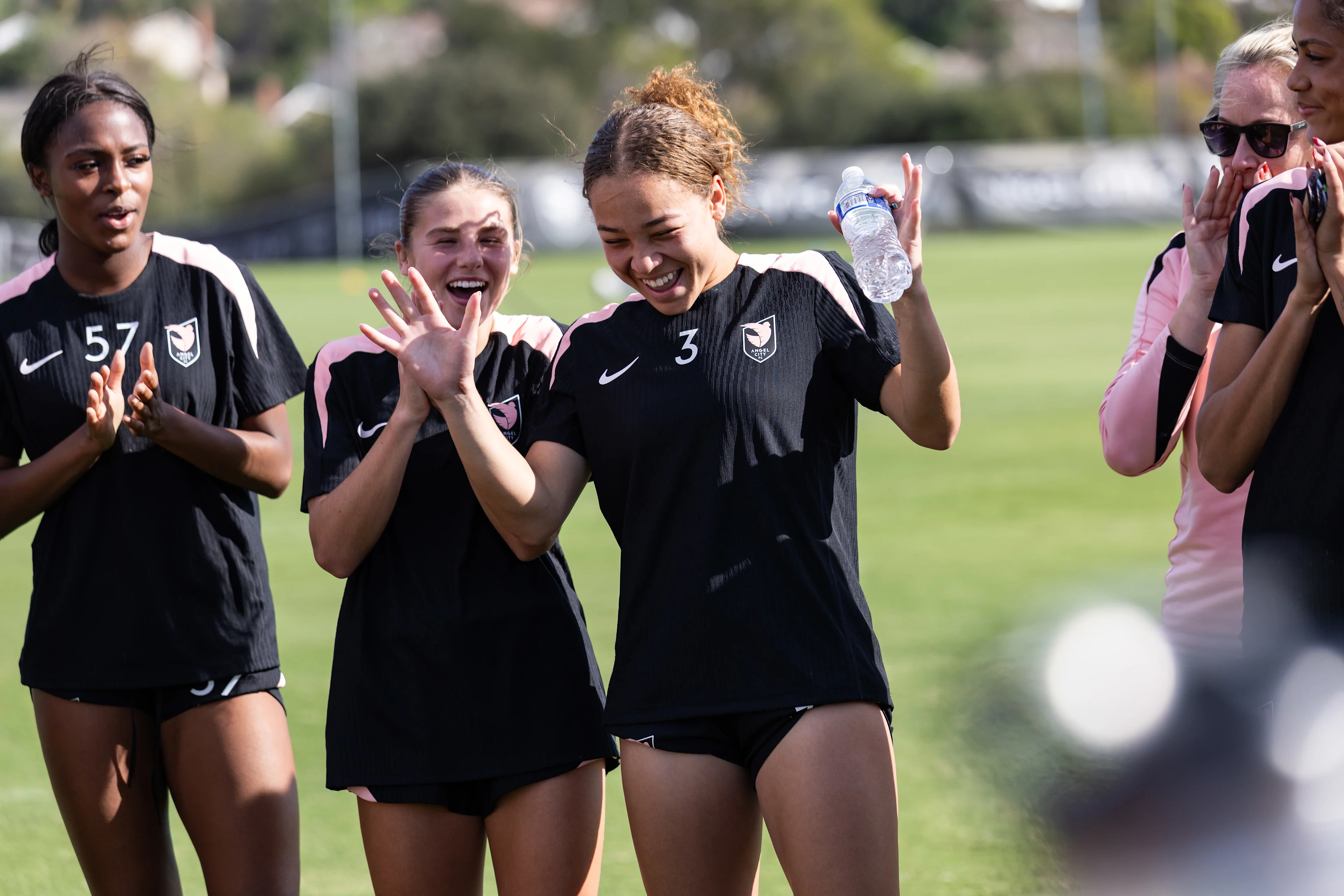 An Angel City FC player in uniform celebrates with her teammates while holding a water bottle after a scrimmage.