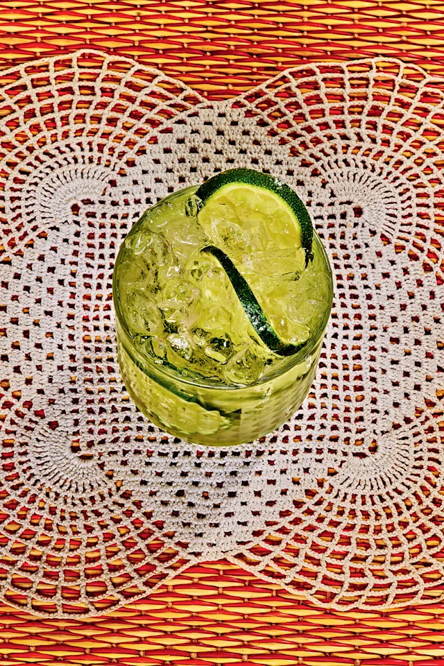 Green cocktail with lime slice and ice in a textured glass on a crocheted doily over red-orange woven placemat.