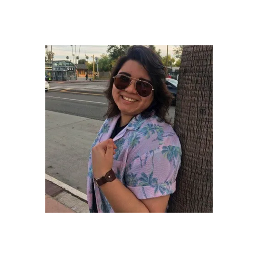 Person wearing sunglasses and a pink palm tree print shirt smiling on a street corner next to a palm tree trunk.