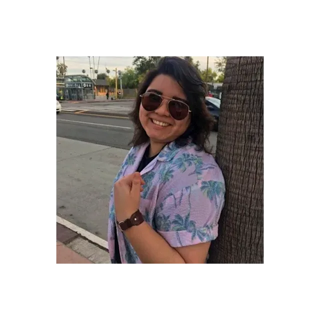 Person wearing sunglasses and a pink palm tree print shirt smiling on a street corner next to a palm tree trunk.