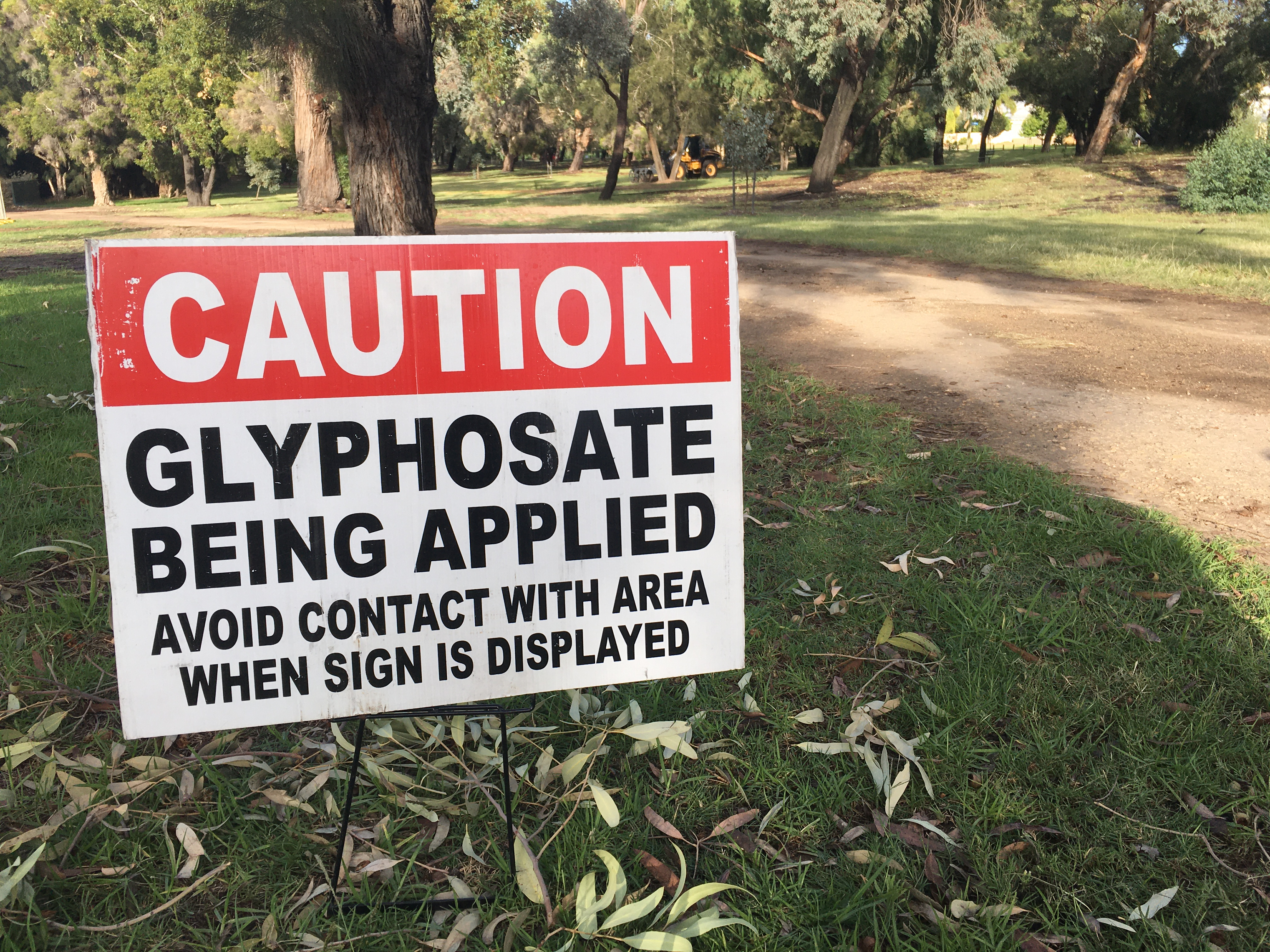 Caution sign warning of glyphosate application in a park with trees and a dirt path, maintenance vehicle visible in background.