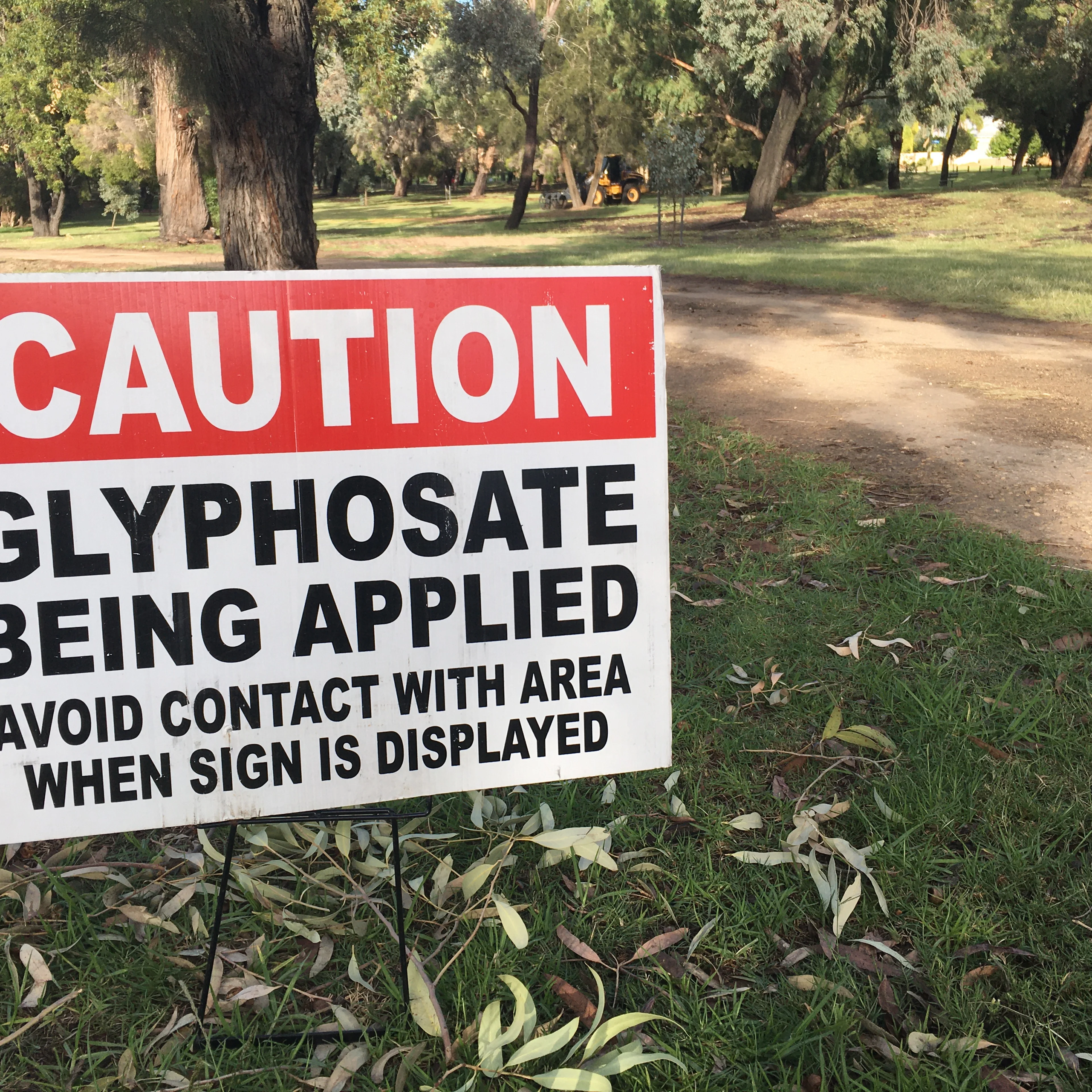 Caution sign warning of glyphosate application in a park with trees and a dirt path, maintenance vehicle visible in background.