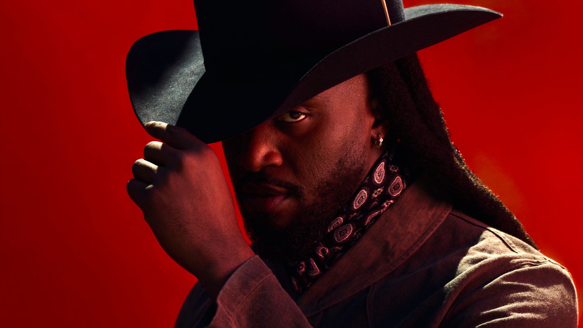 A man in a black cowboy hat and bandana gazes intensely against a bold red background, with dramatic lighting.
