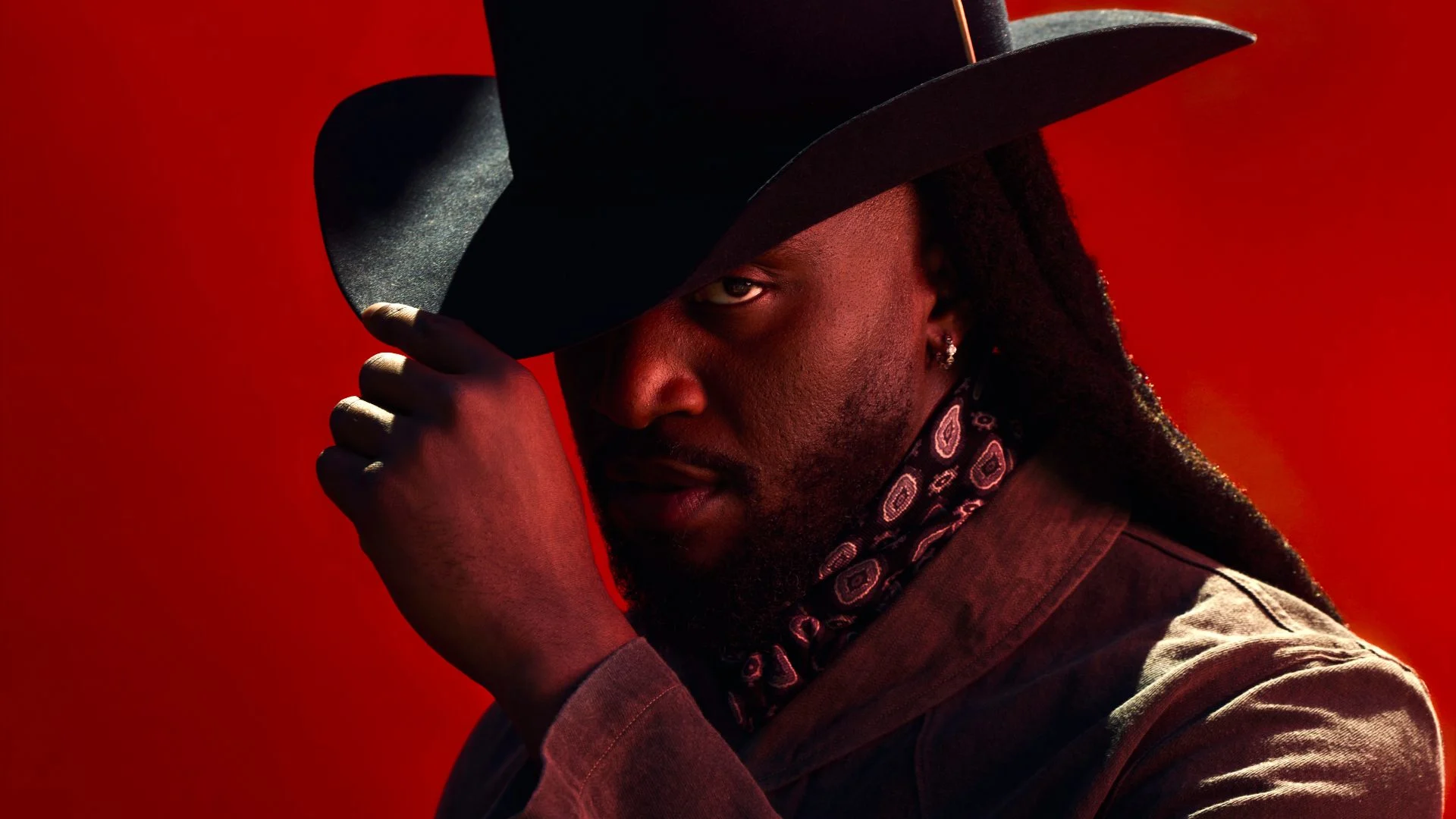 A man in a black cowboy hat and bandana gazes intensely against a bold red background, with dramatic lighting.