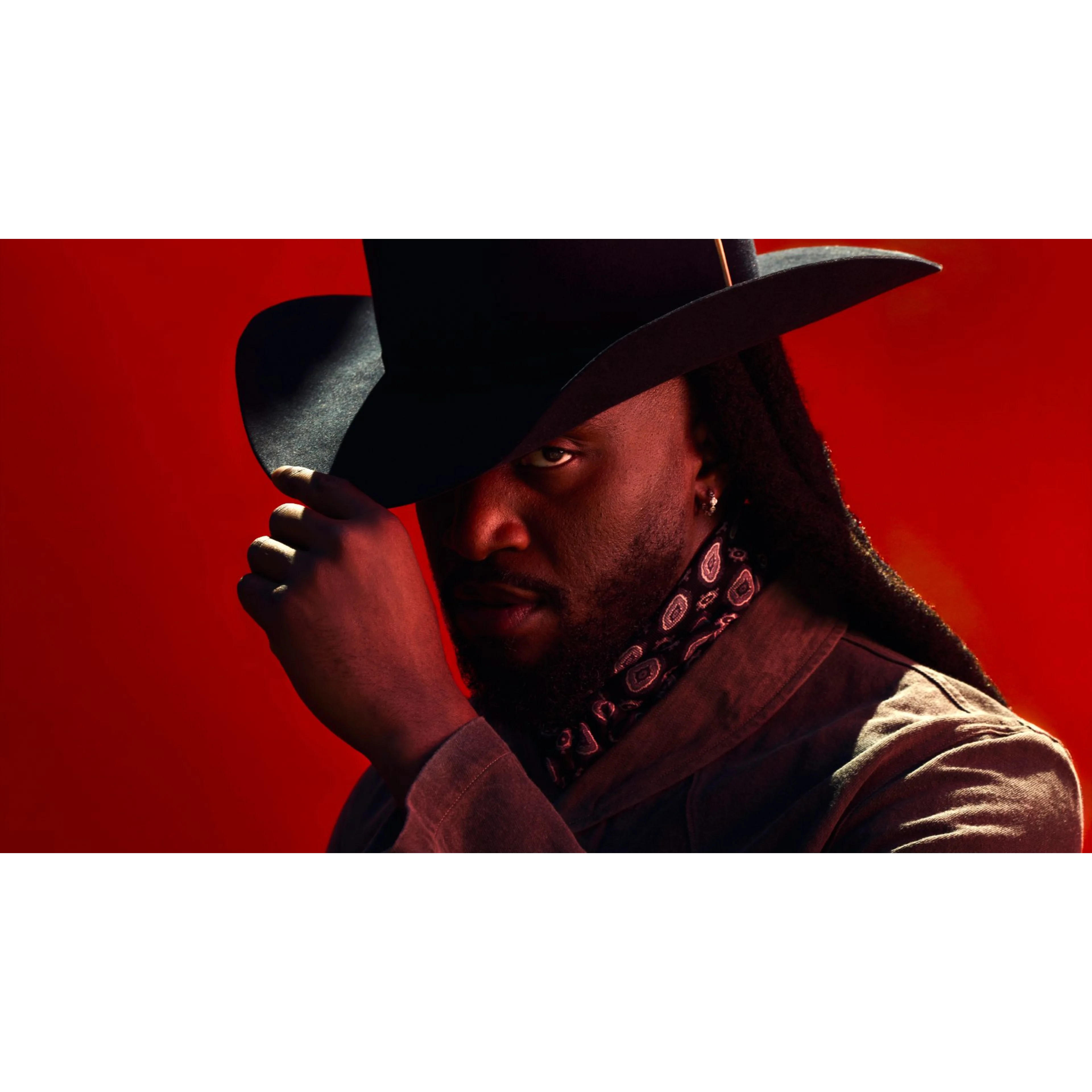 A man in a black cowboy hat and bandana gazes intensely against a bold red background, with dramatic lighting.