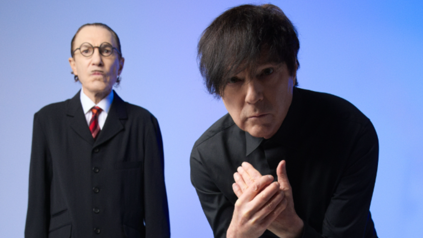 Two musicians in black suits against a blue background, one with glasses, the other with dark hair clasping hands.