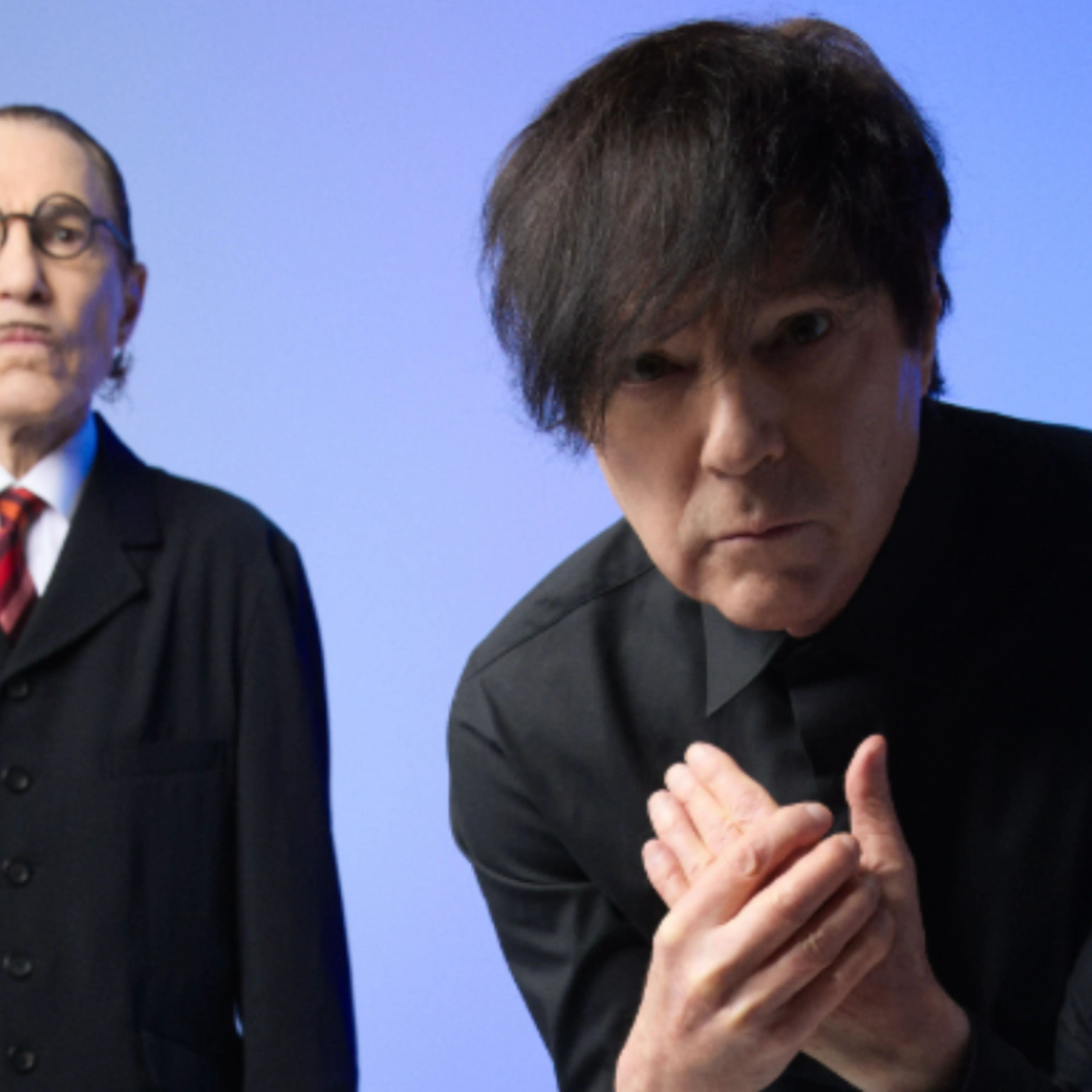 Two musicians in black suits against a blue background, one with glasses, the other with dark hair clasping hands.
