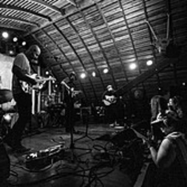 A black and white photo of a band performing on stage in a venue with an arched ceiling and audience silhouettes.