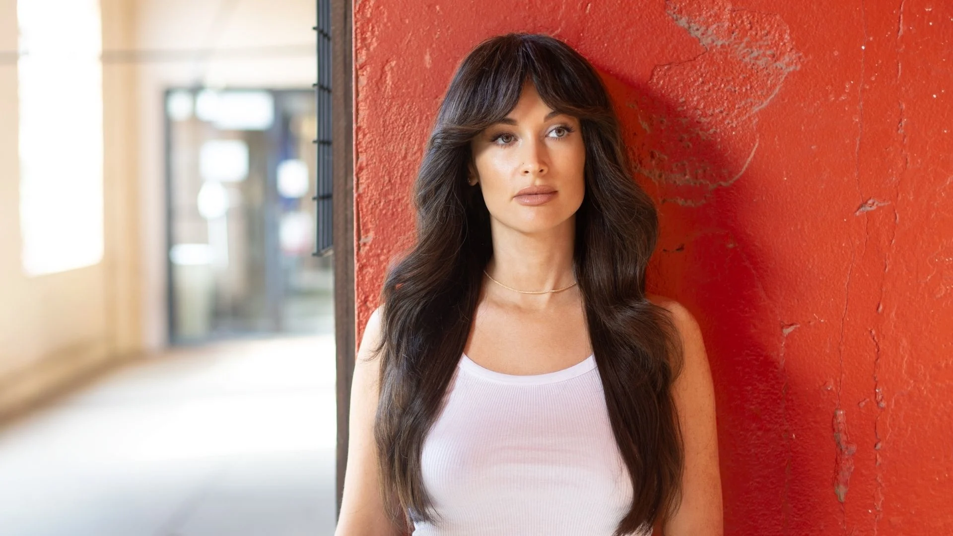 A woman with long dark wavy hair and bangs leans against a red wall, wearing a white tank top and gold necklace.