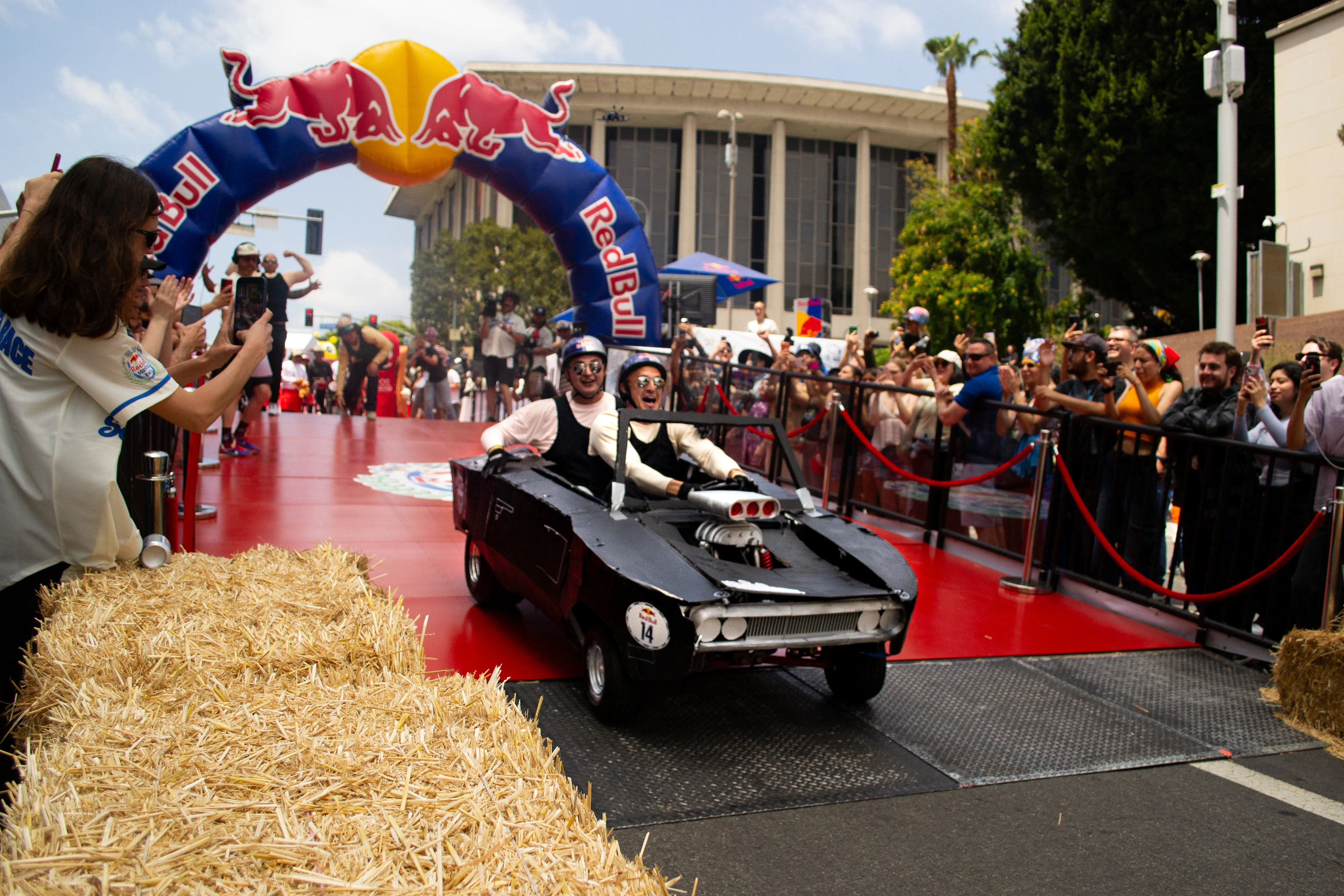 Two men in tank tops and white, long-sleeved undershirts ride in a handmade soapbox car as they launch off the starting platform surrounded by a crowd.
