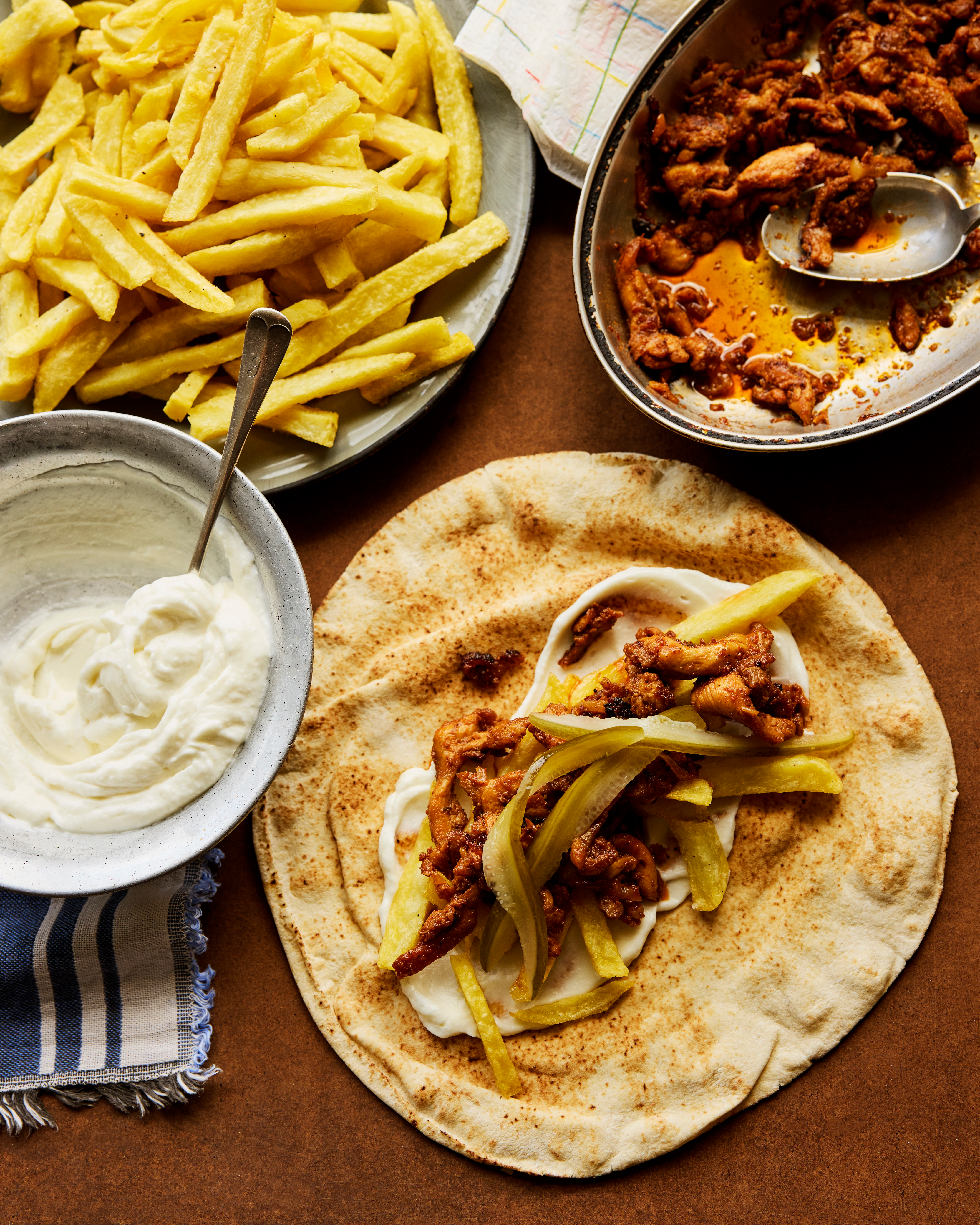 A flatbread topped with spiced meat and pickles, served alongside french fries, creamy sauce, and a pan of seasoned chicken.
