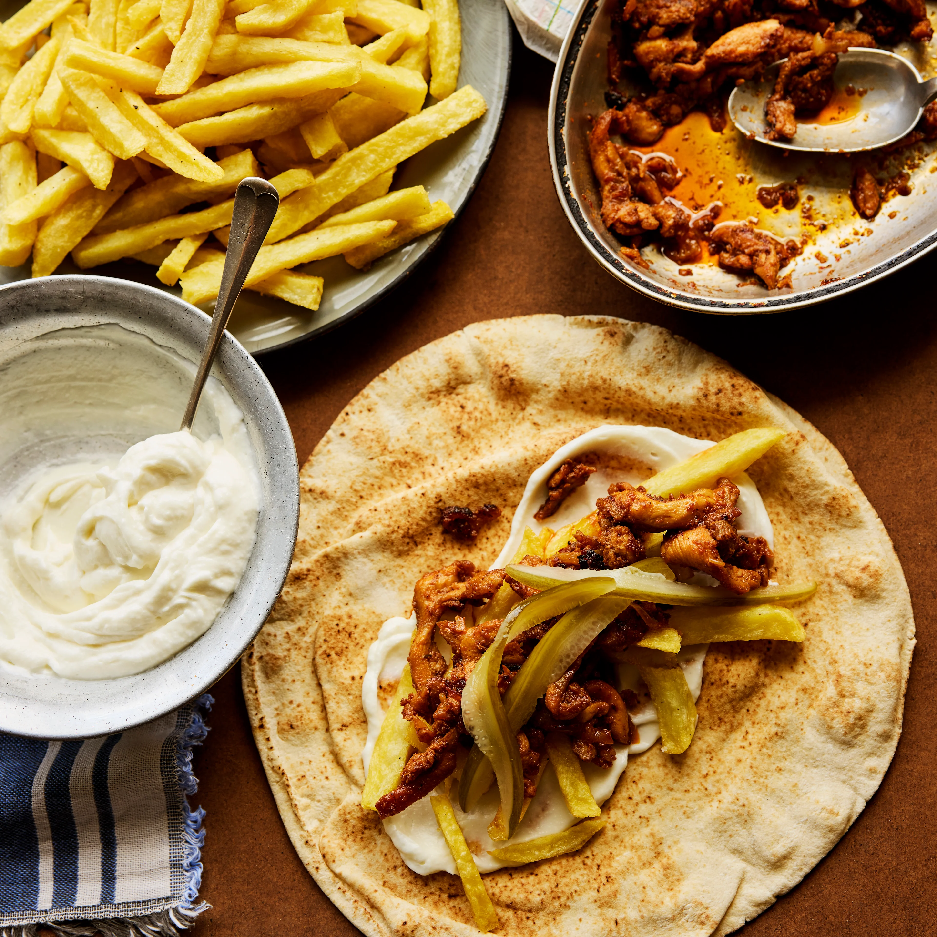 A flatbread topped with spiced meat and pickles, served alongside french fries, creamy sauce, and a pan of seasoned chicken.
