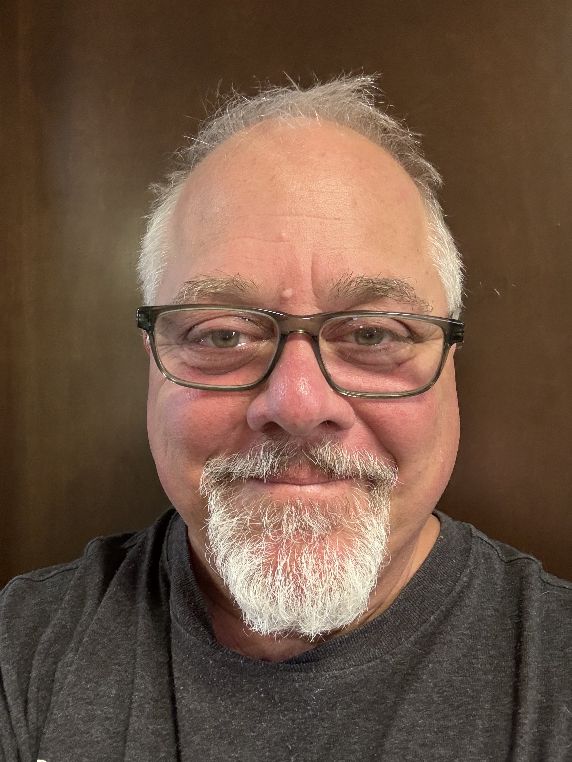 A middle-aged man with a white goatee, glasses, and graying hair smiles at the camera against a brown background.