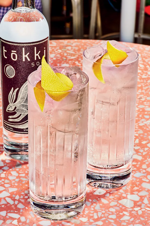 Two pink cocktails with ice and lemon garnish next to a bottle of Tōkki spirit on a coral-colored table.