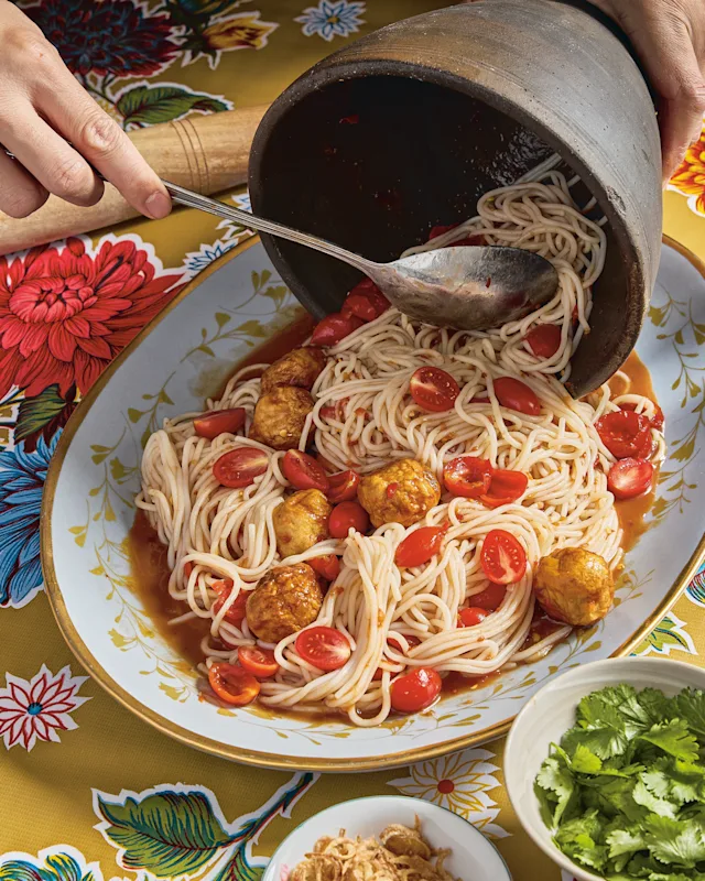 Hands pouring rich tomato broth over noodles, meatballs, and cherry tomatoes in a floral bowl on a yellow tablecloth.