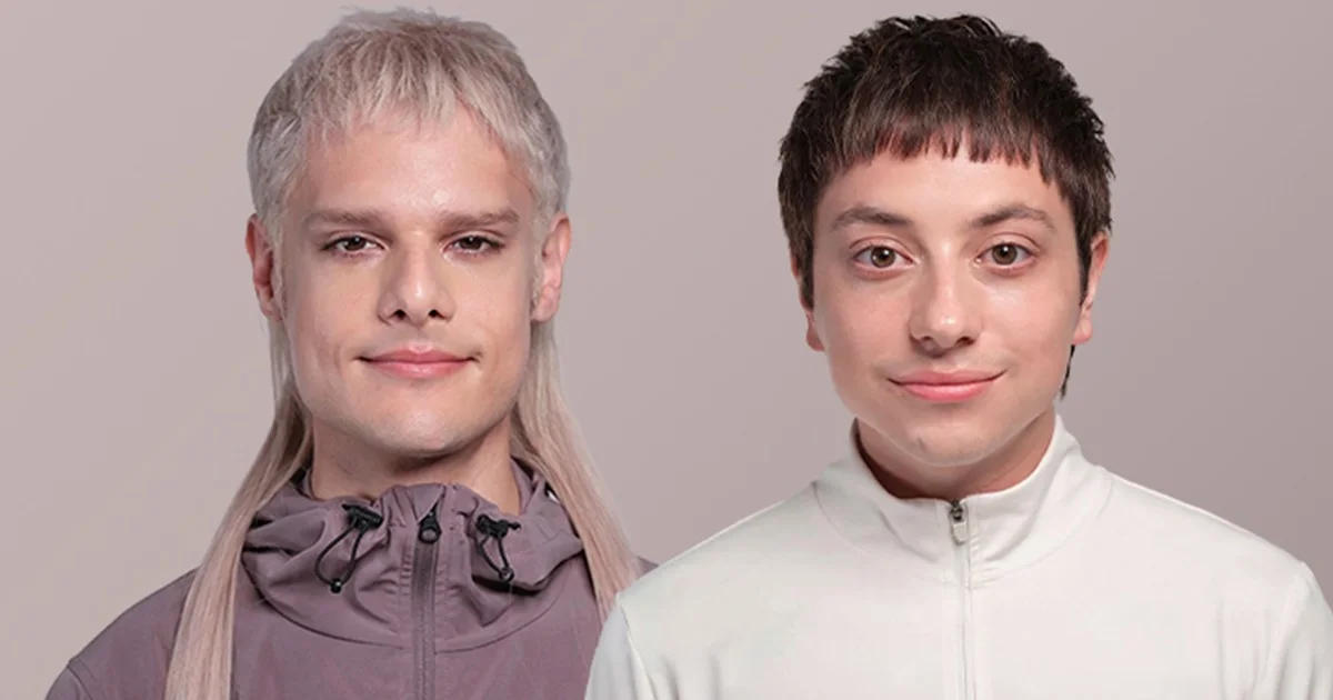 Two young men posing together, one with long platinum blonde hair in a mauve jacket, the other with short dark hair in a white jacket.