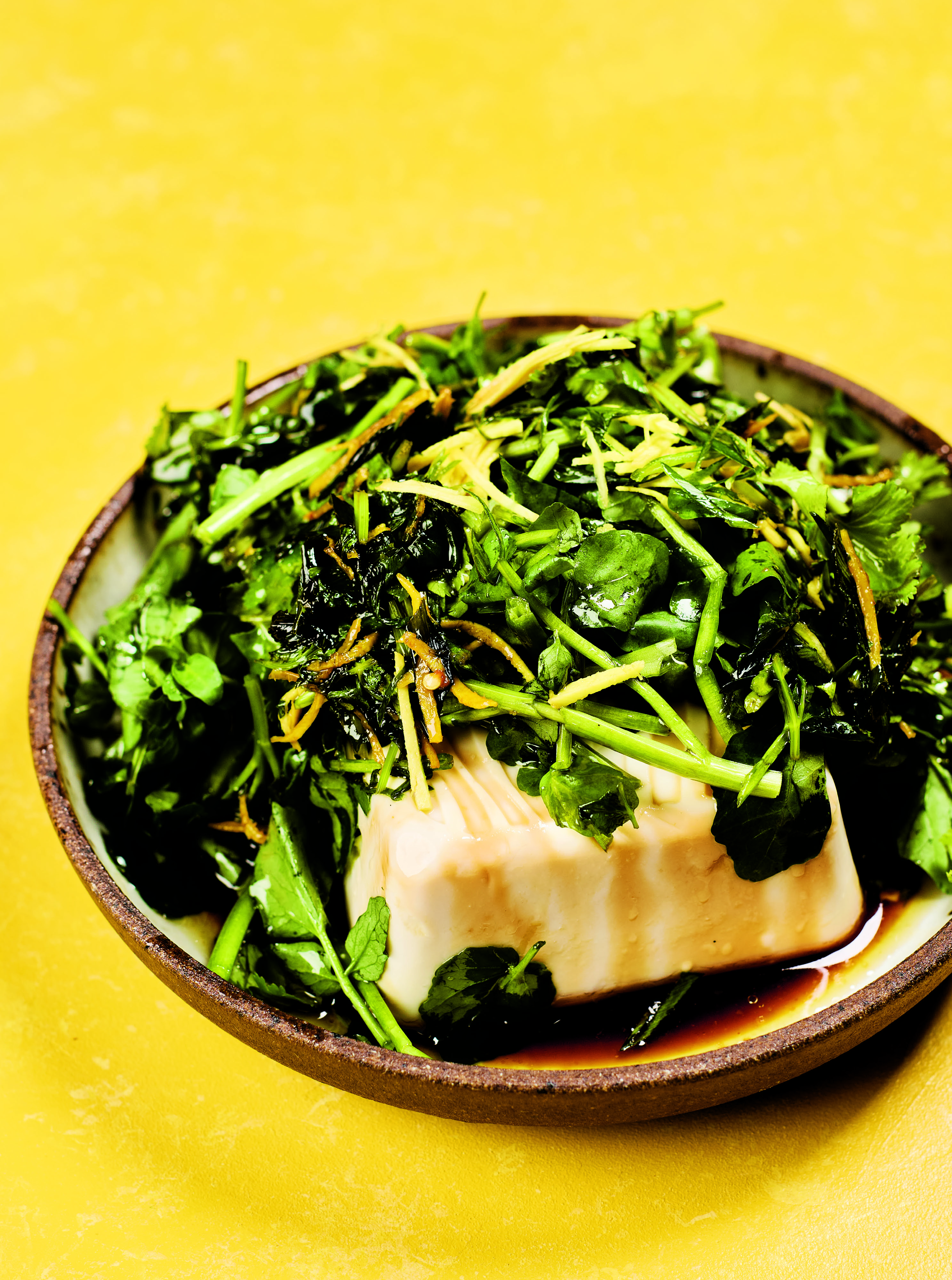 Silken tofu topped with fresh green herbs and soy sauce in a ceramic bowl against a bright yellow background.