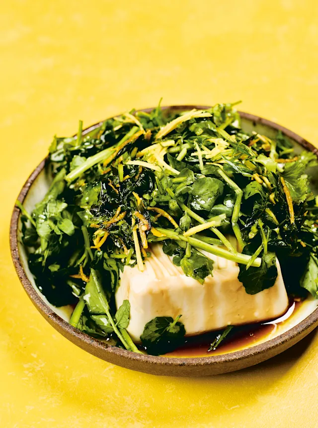 Silken tofu topped with fresh green herbs and soy sauce in a ceramic bowl against a bright yellow background.