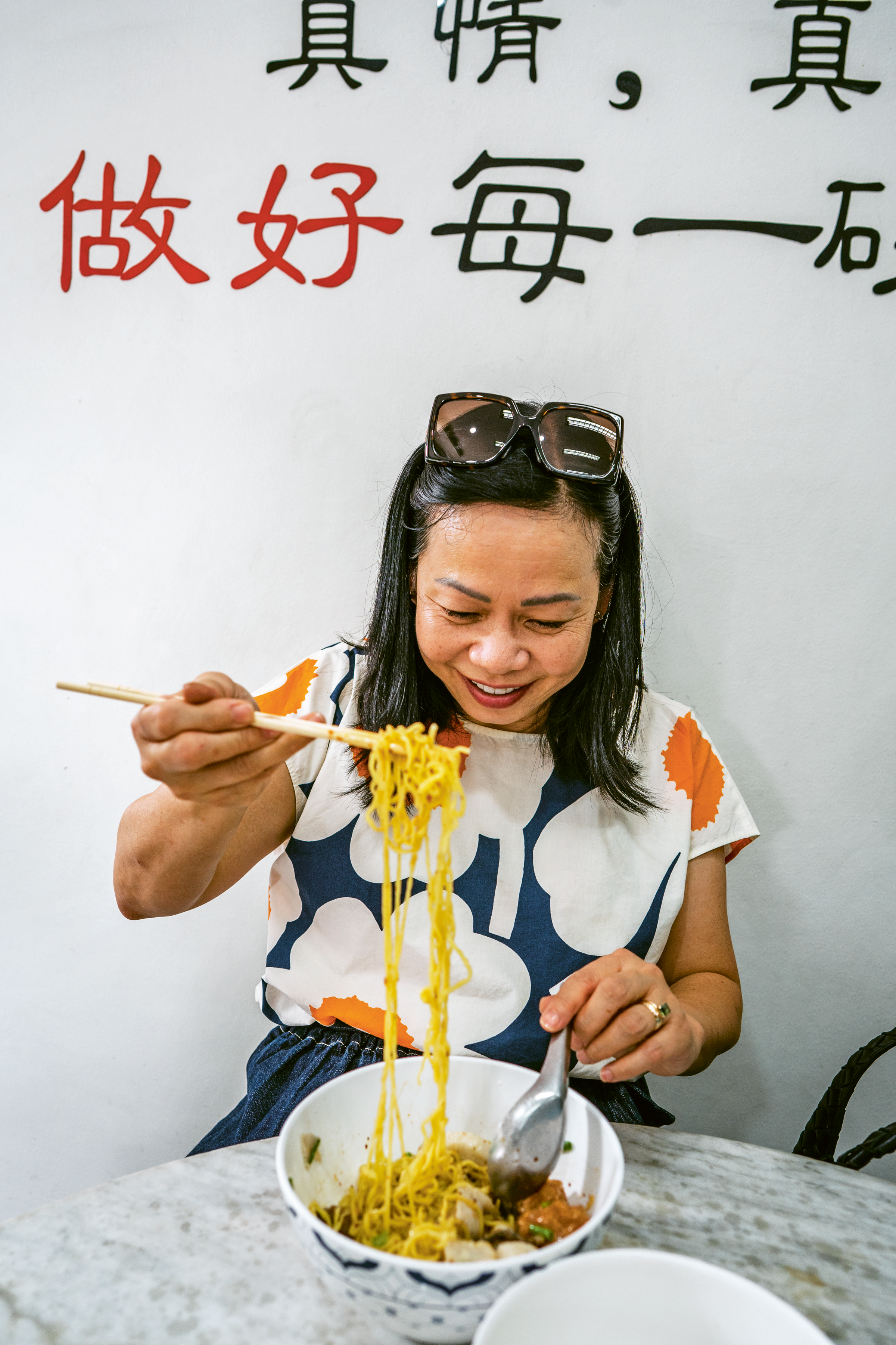 A smiling woman uses chopsticks to lift noodles from a bowl at a restaurant with Chinese characters on the wall behind her.