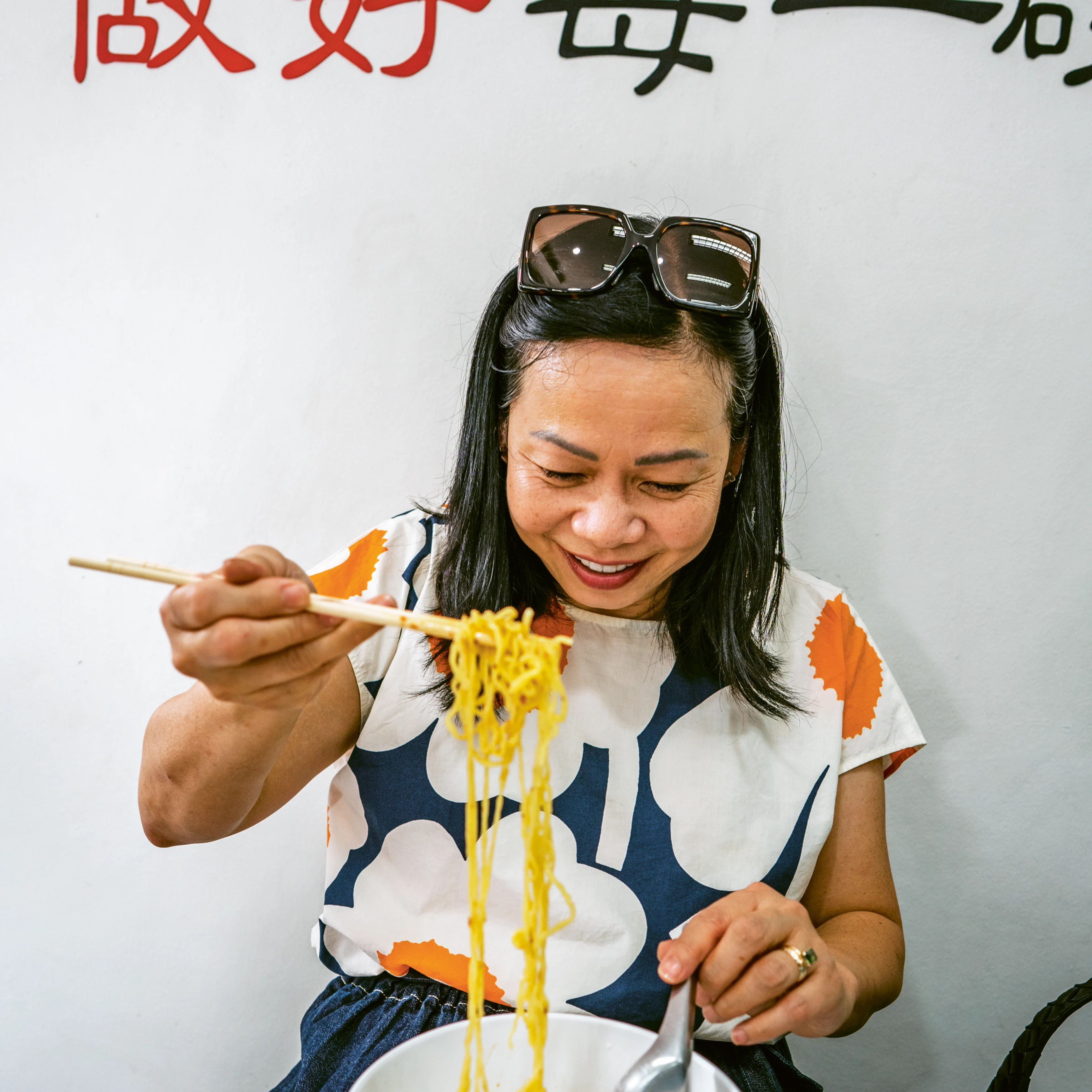 A smiling woman uses chopsticks to lift noodles from a bowl at a restaurant with Chinese characters on the wall behind her.