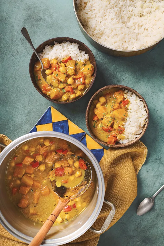 A pot of chickpea and vegetable curry with a wooden ladle, served in two bowls with white rice on a teal surface.