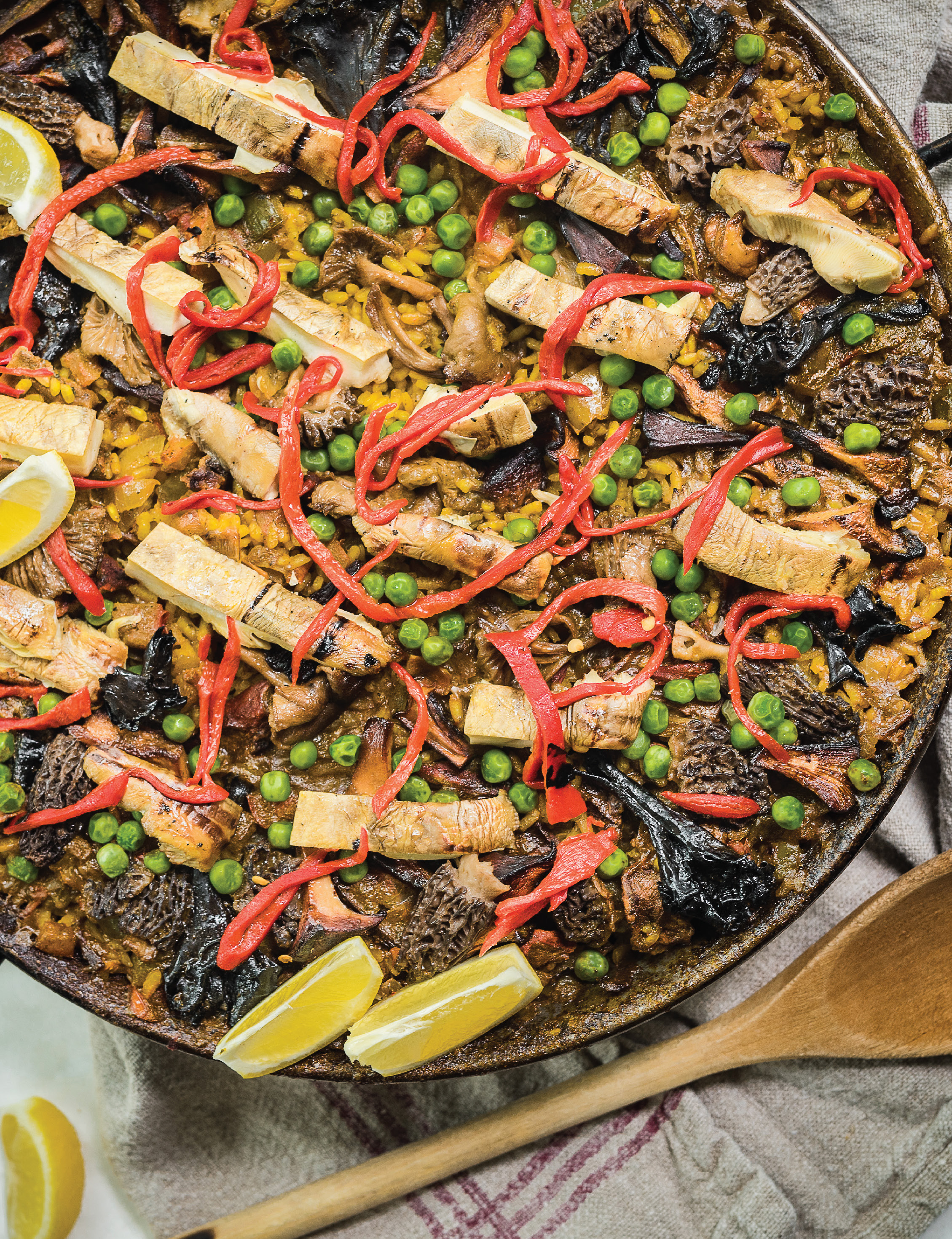 Traditional Spanish paella with saffron rice, green peas, red pepper strips, mushrooms, and lemon wedges in a round pan.