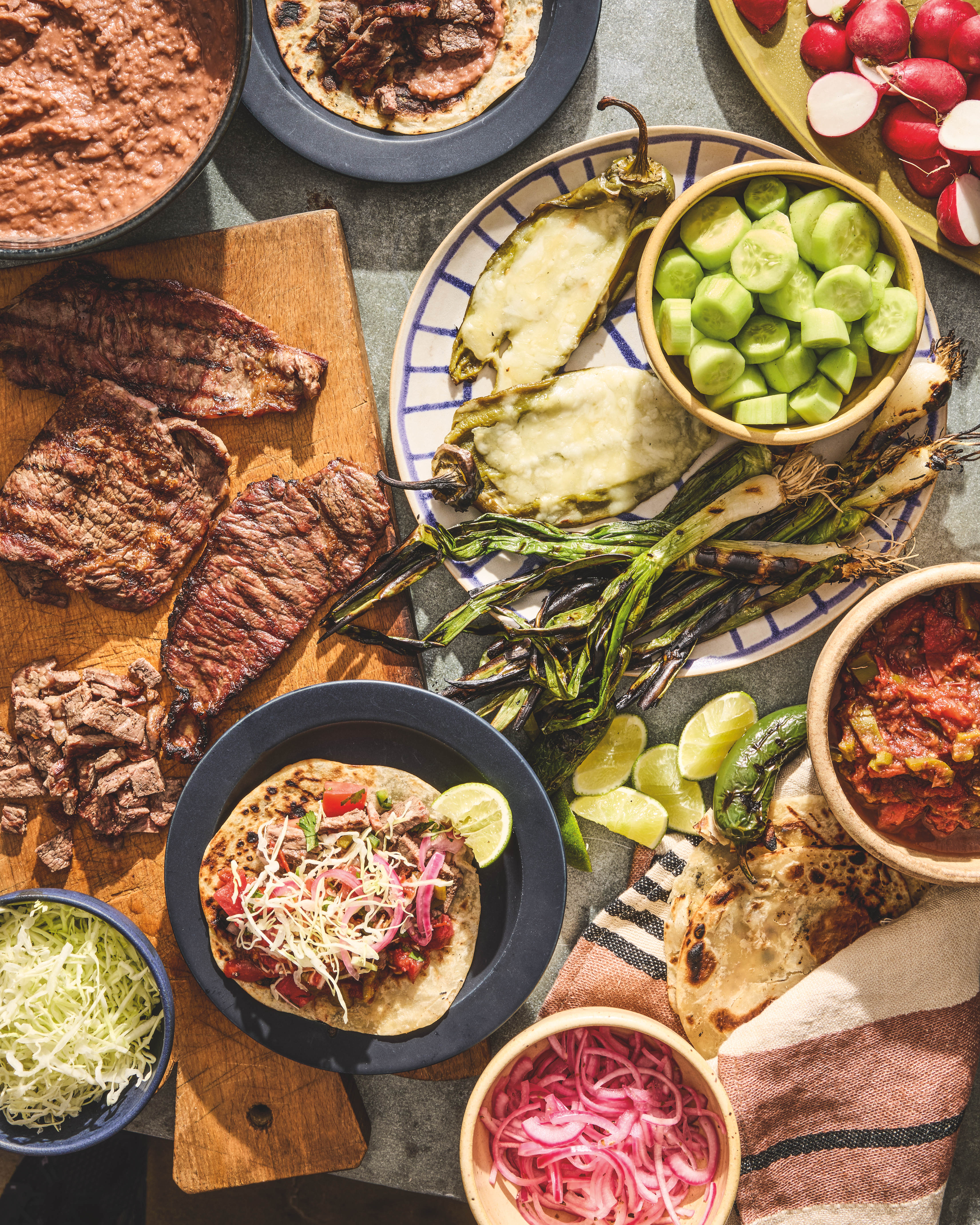 A spread of Mexican food including grilled steak, tacos, roasted peppers, cucumber, pickled onions, salsa, and refried beans.